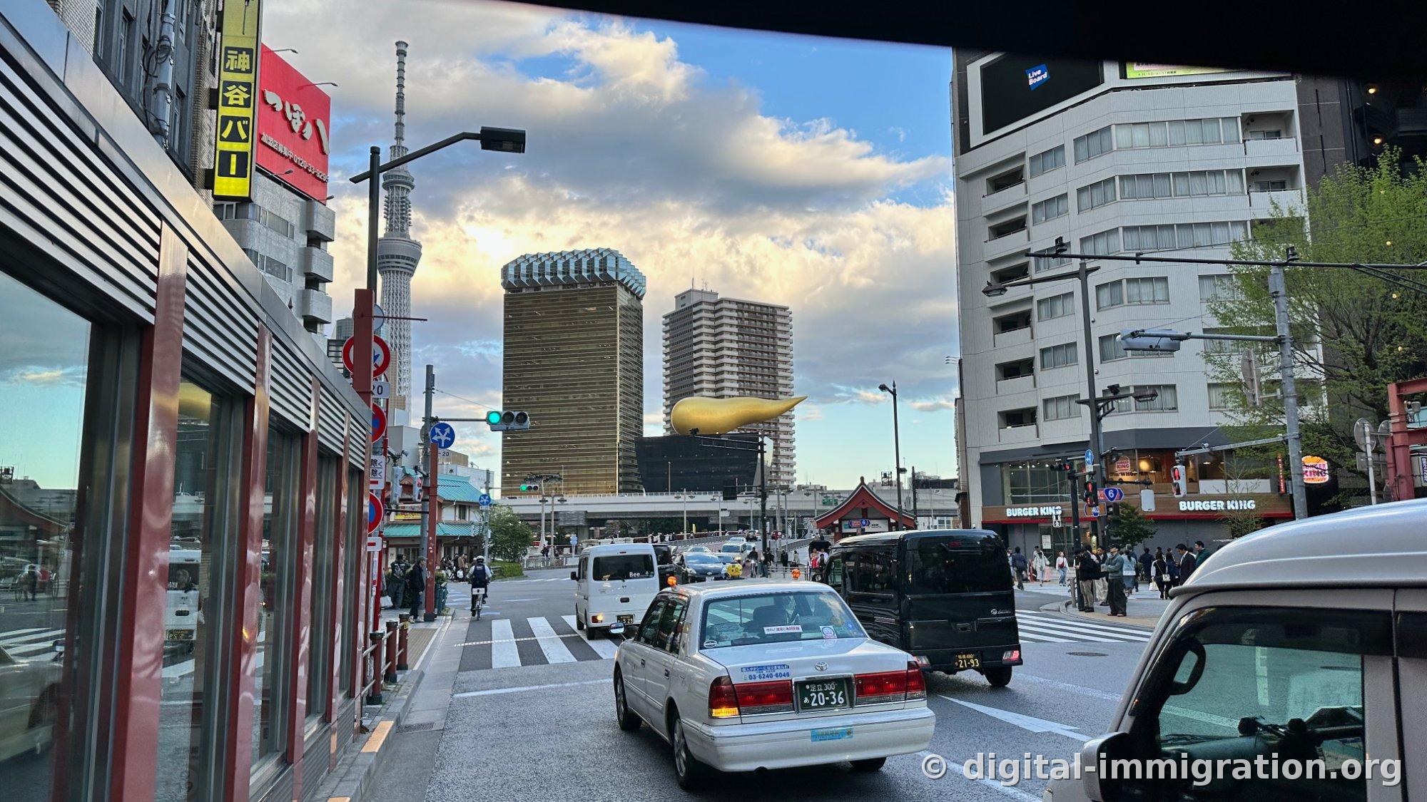 The view from Azumabashi Intersection at sunset. Tokyo Skytree on the left, the Asahi brewery building and golden flame sculpture in the center, a Burger King sign and white taxi on the right.