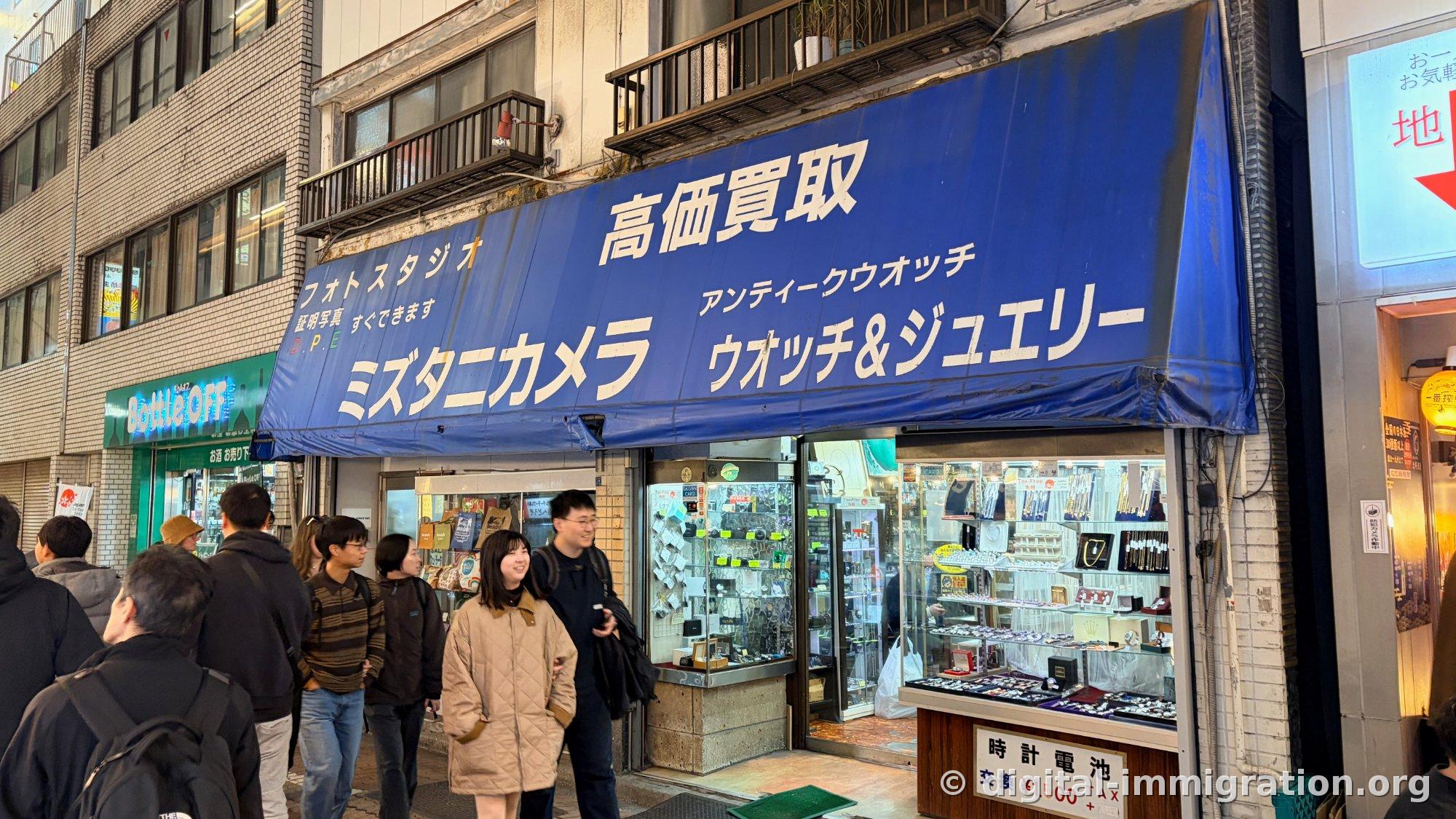 Une foule passant devant un magasin avec un auvent bleu inscrit « 高価買取 ミズタニカメラ ウオッチ&ジュエリー », des montres et accessoires exposés dans la vitrine
