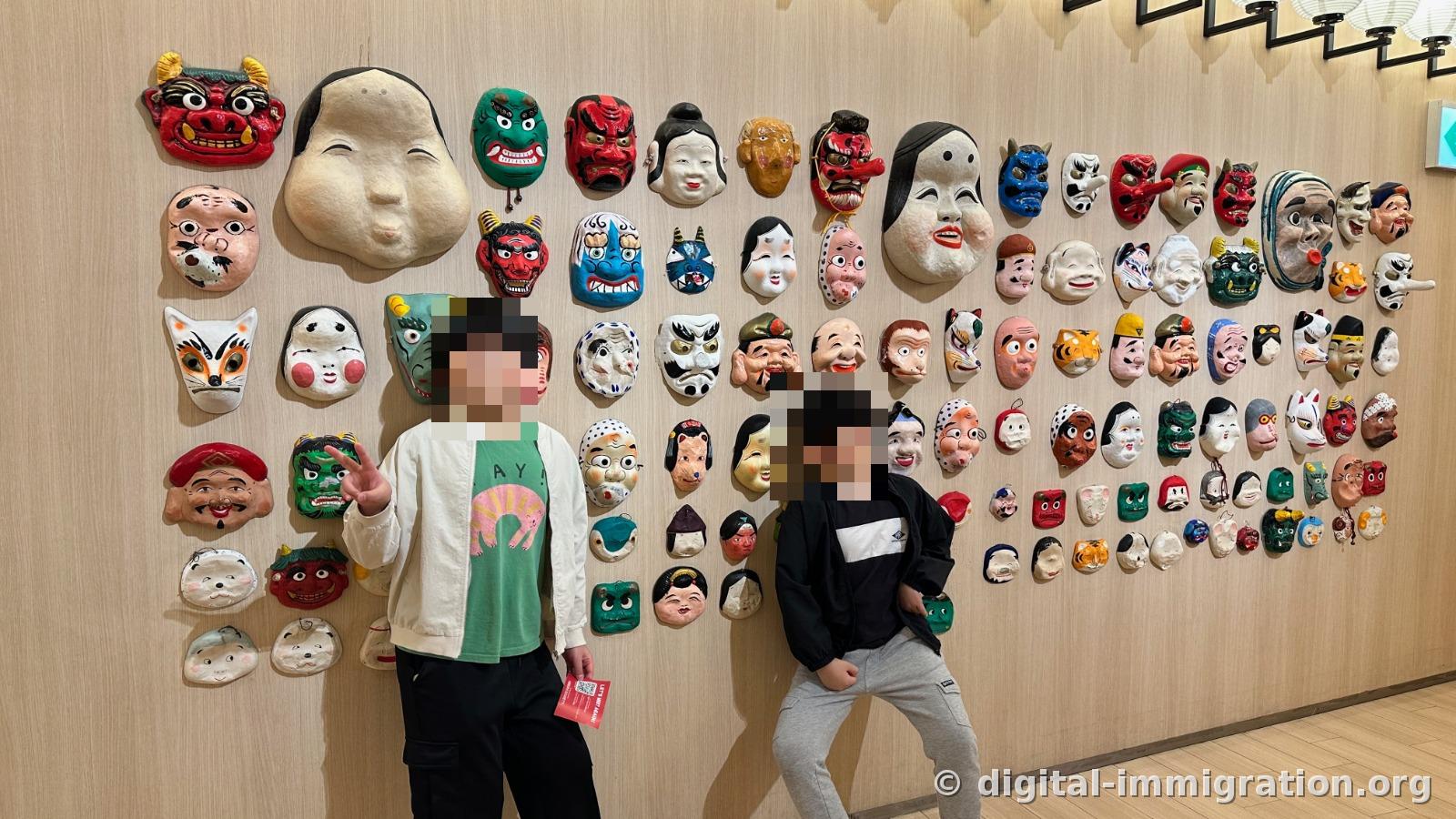 Deux enfants posant devant l'exposition de masques traditionnels japonais qui couvre entièrement le mur. Des masques rouges de démons, des masques blancs Okame, des masques de renards, des tengus et autres sont disposés par taille, près d'une centaine