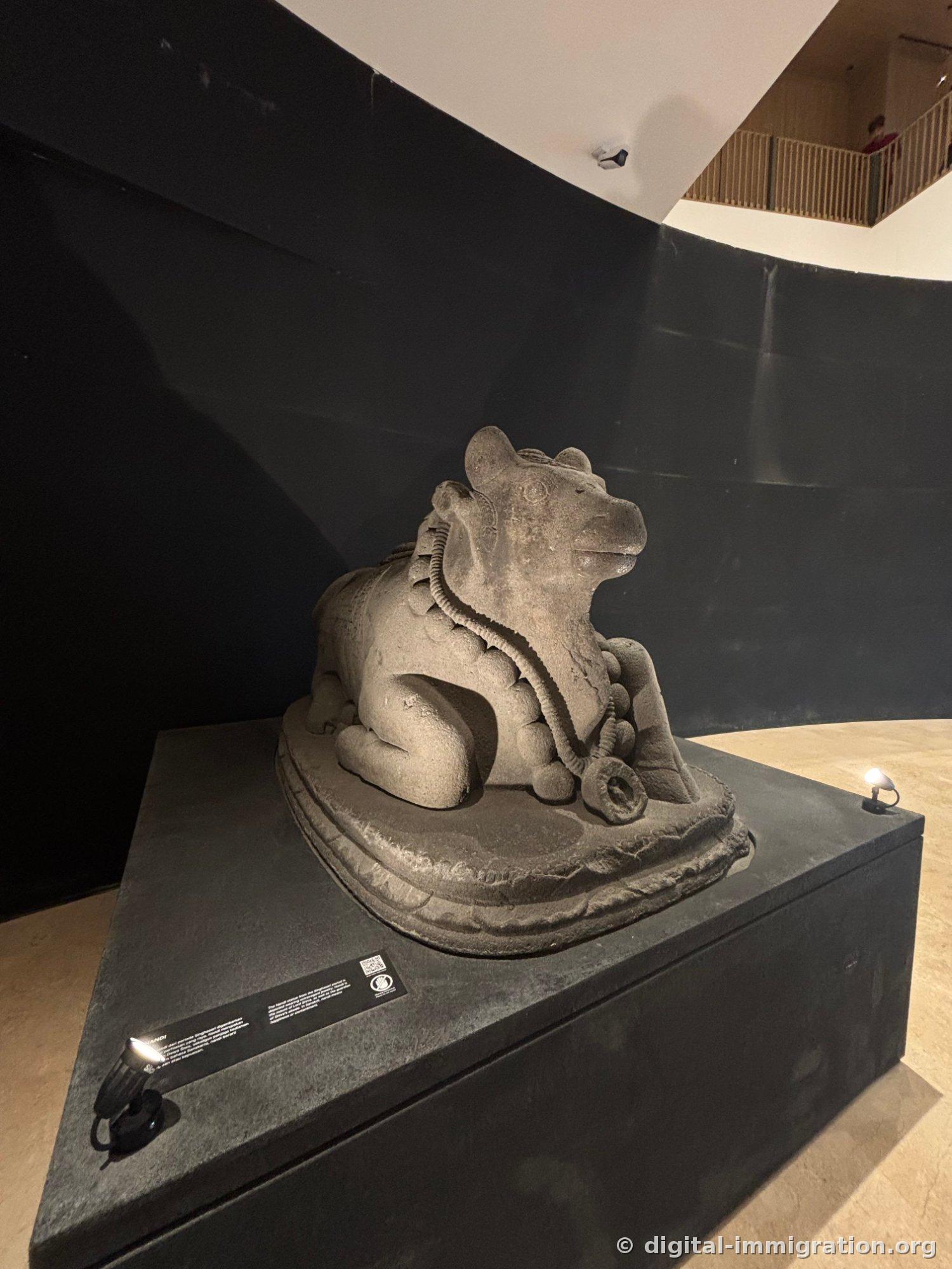 A carved stone bull (Nandi) crouching on a pedestal against a curved black wall, with a bell ornament on its neck