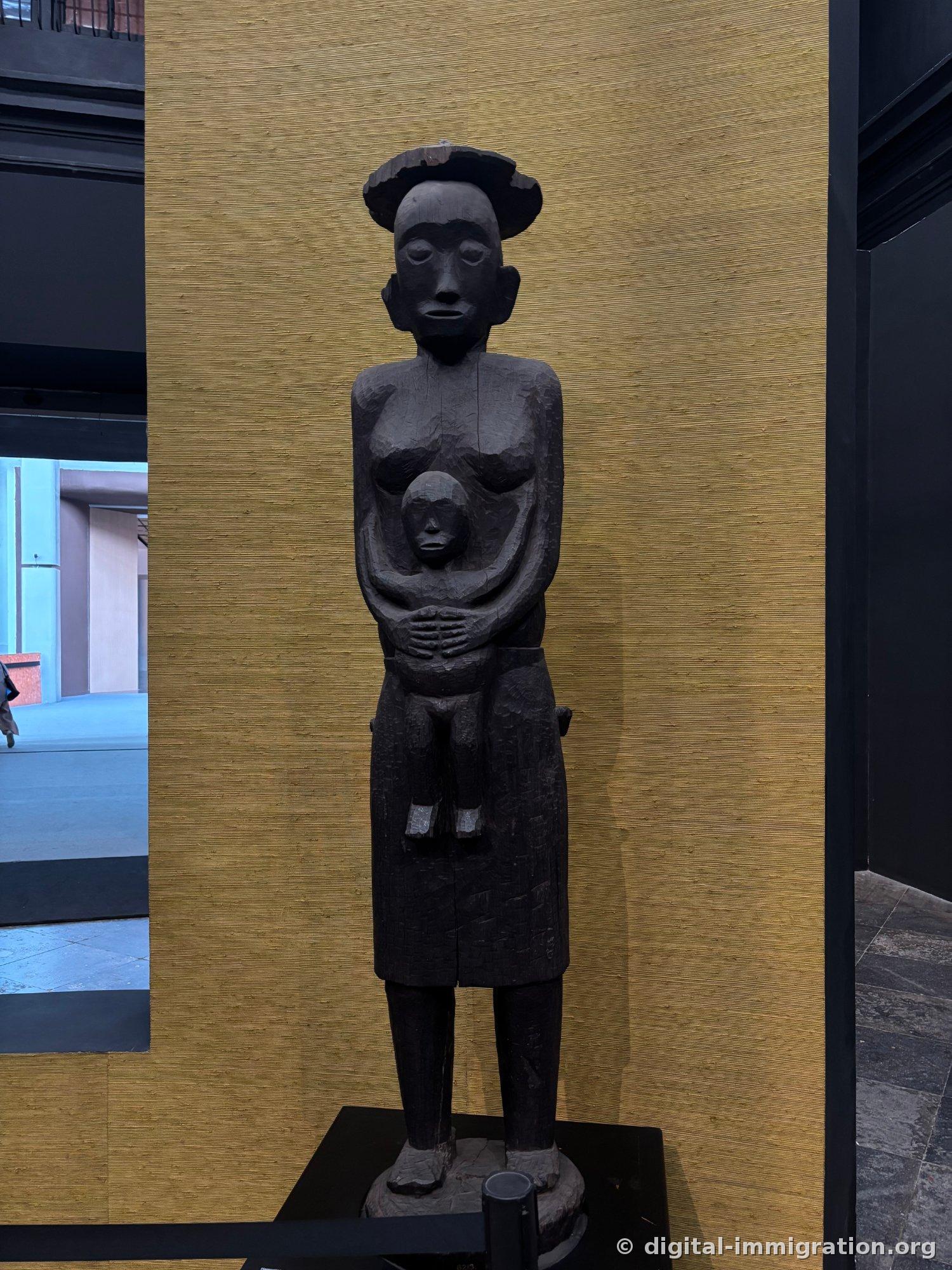 A black wooden ancestral figure standing before a golden straw-textured wall, wearing a broad-brimmed headdress and holding a child
