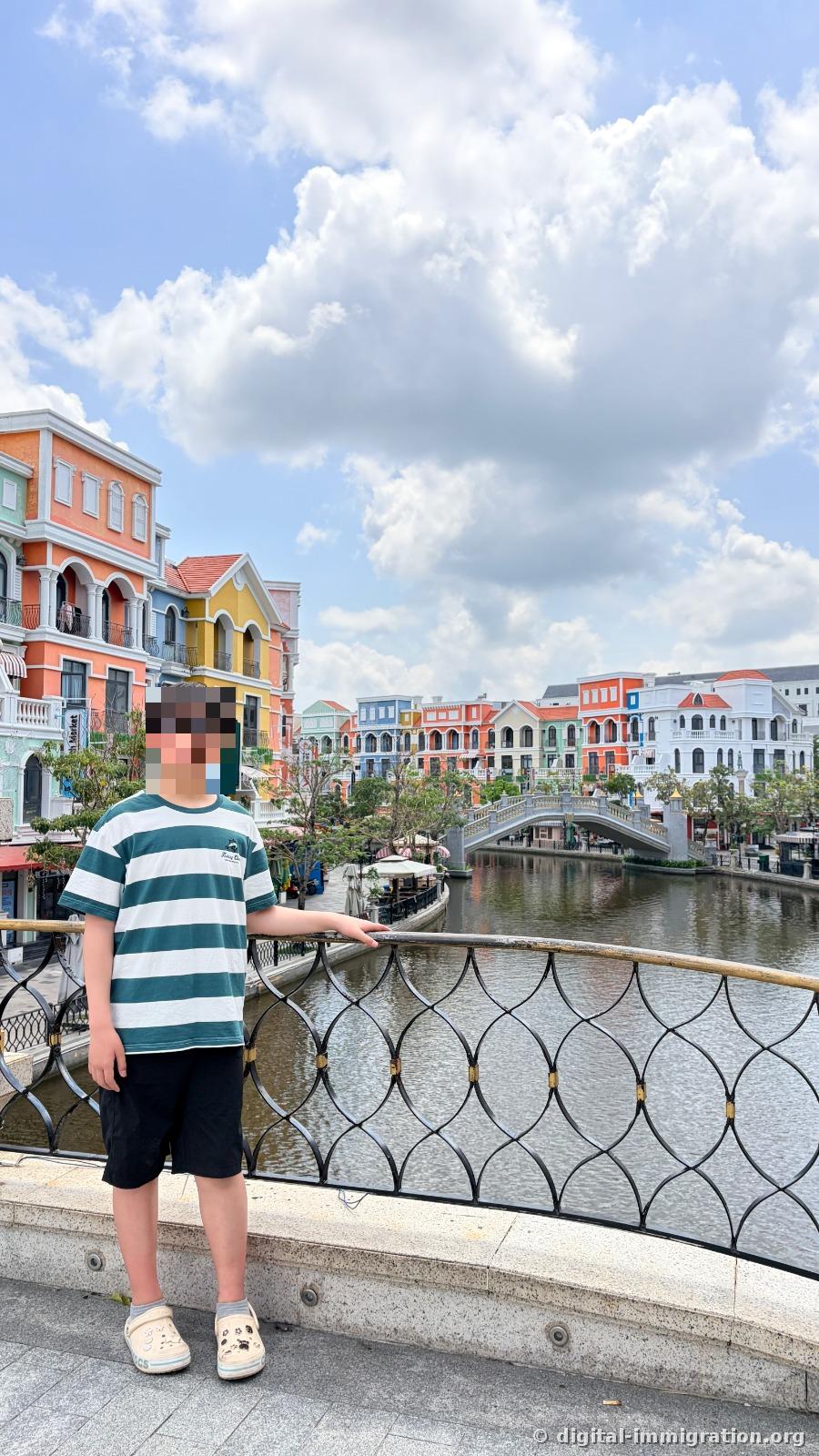 A boy leaning against a canal railing under a cloudy blue sky, with a white arch bridge visible on the far bank