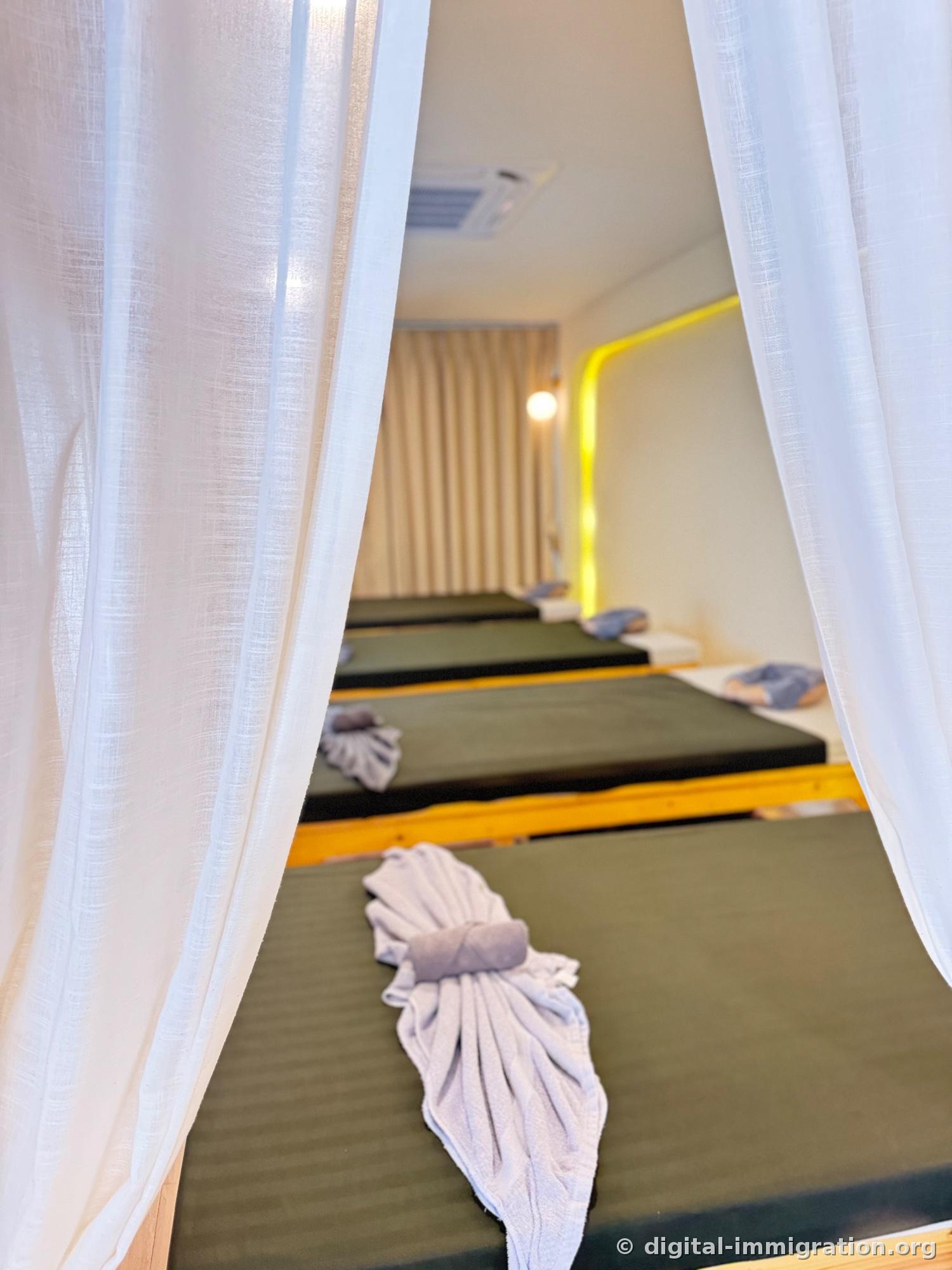 Shared massage room with white linen curtains parted to reveal four beds with green mats lined up in a row, folded towels on each bed, ceiling air conditioner visible