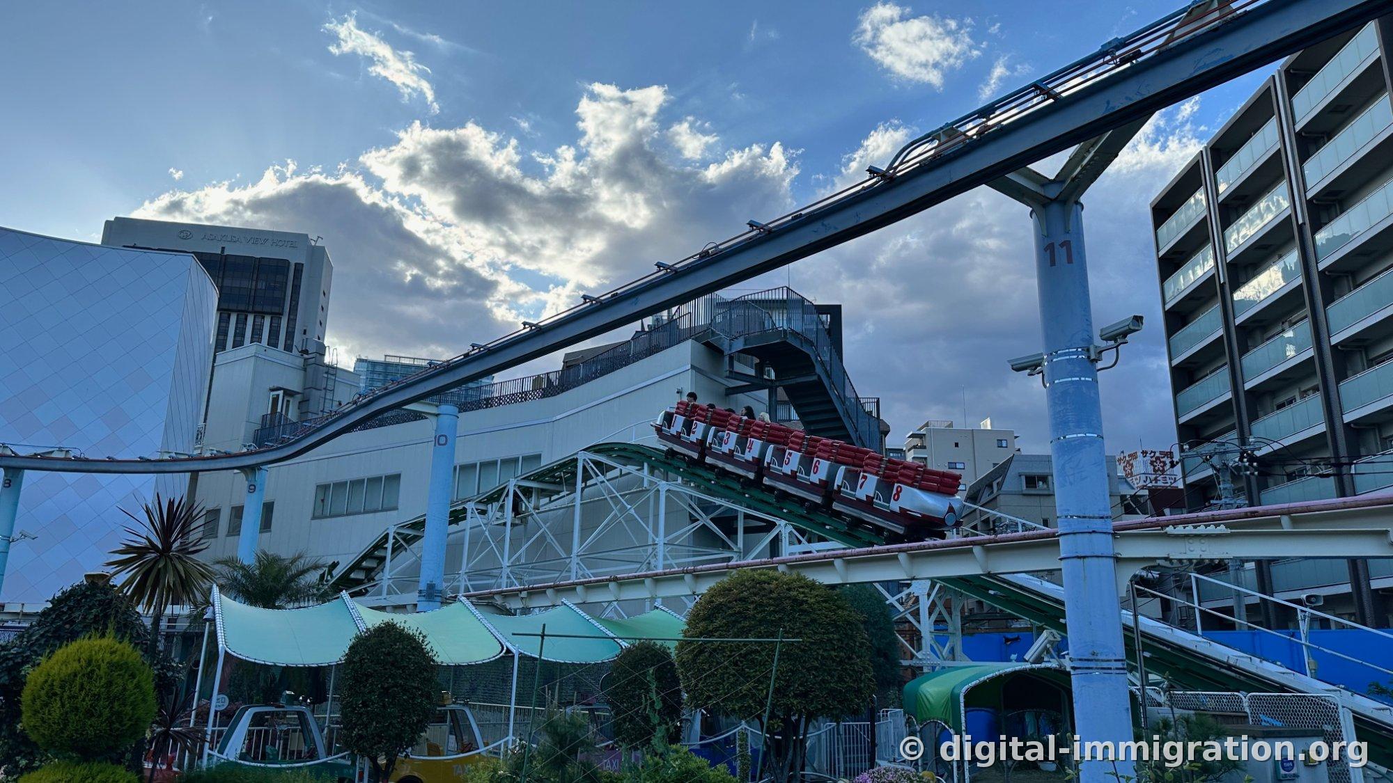 Hanayashiki roller coaster rails and 8-car train under cloudy sky, with ASAKUSA VIEW HOTEL building behind