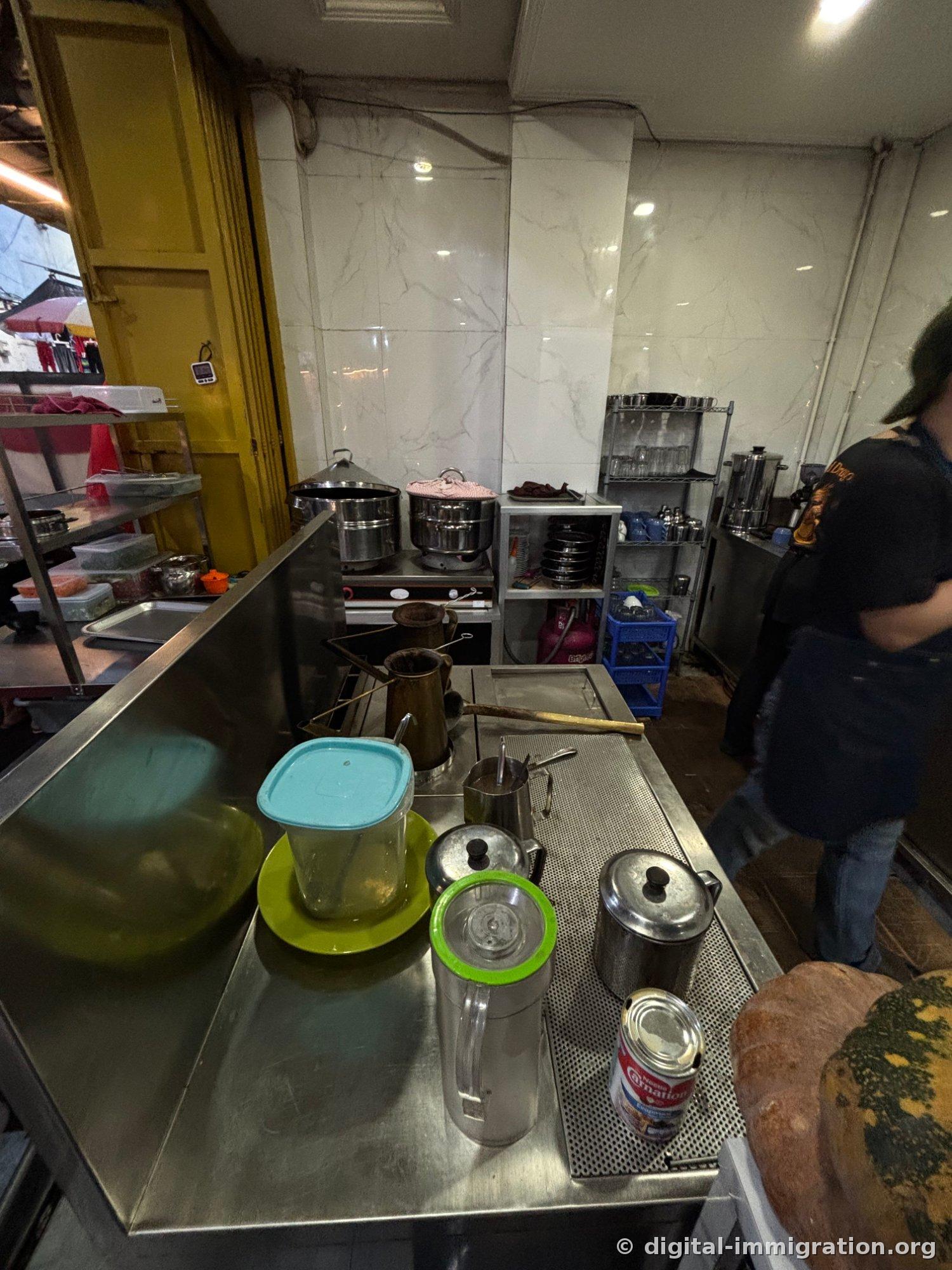 Derrière le comptoir en acier inoxydable, deux paniers vapeur sont sur le feu et une théière en cuivre, une cafetière, une boîte de lait condensé et une courge sont disposés. Un employé en tablier se tient devant