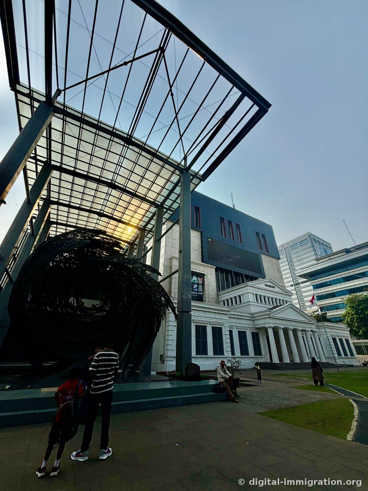 The black spiral sculpture in the courtyard at dusk, with the white neoclassical main building beyond the glass canopy and the gray new wing behind