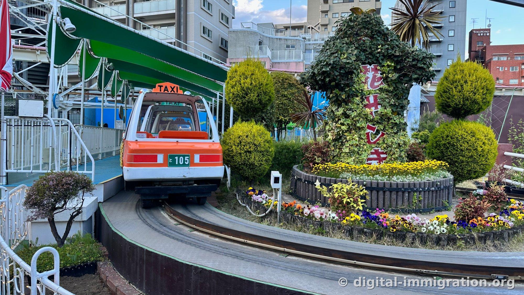 Orange TAXI ride running on rails under green canopy, 花やしき sign covered with ivy, and flower bed with pansies