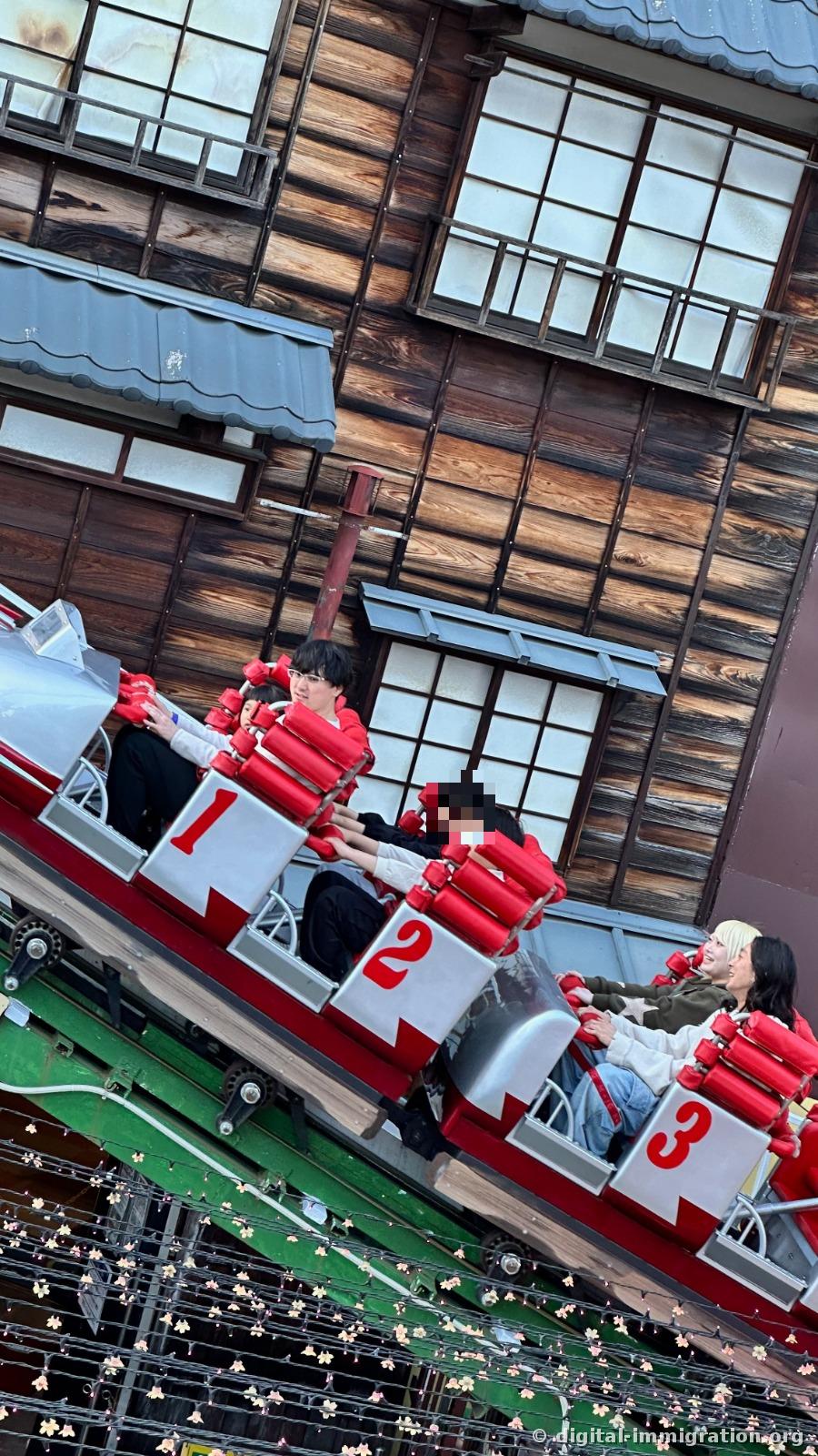 Roller coaster car climbing diagonally with a wooden building wall as backdrop, passengers in seats 1, 2, 3, and cherry blossom decoration lights hanging below