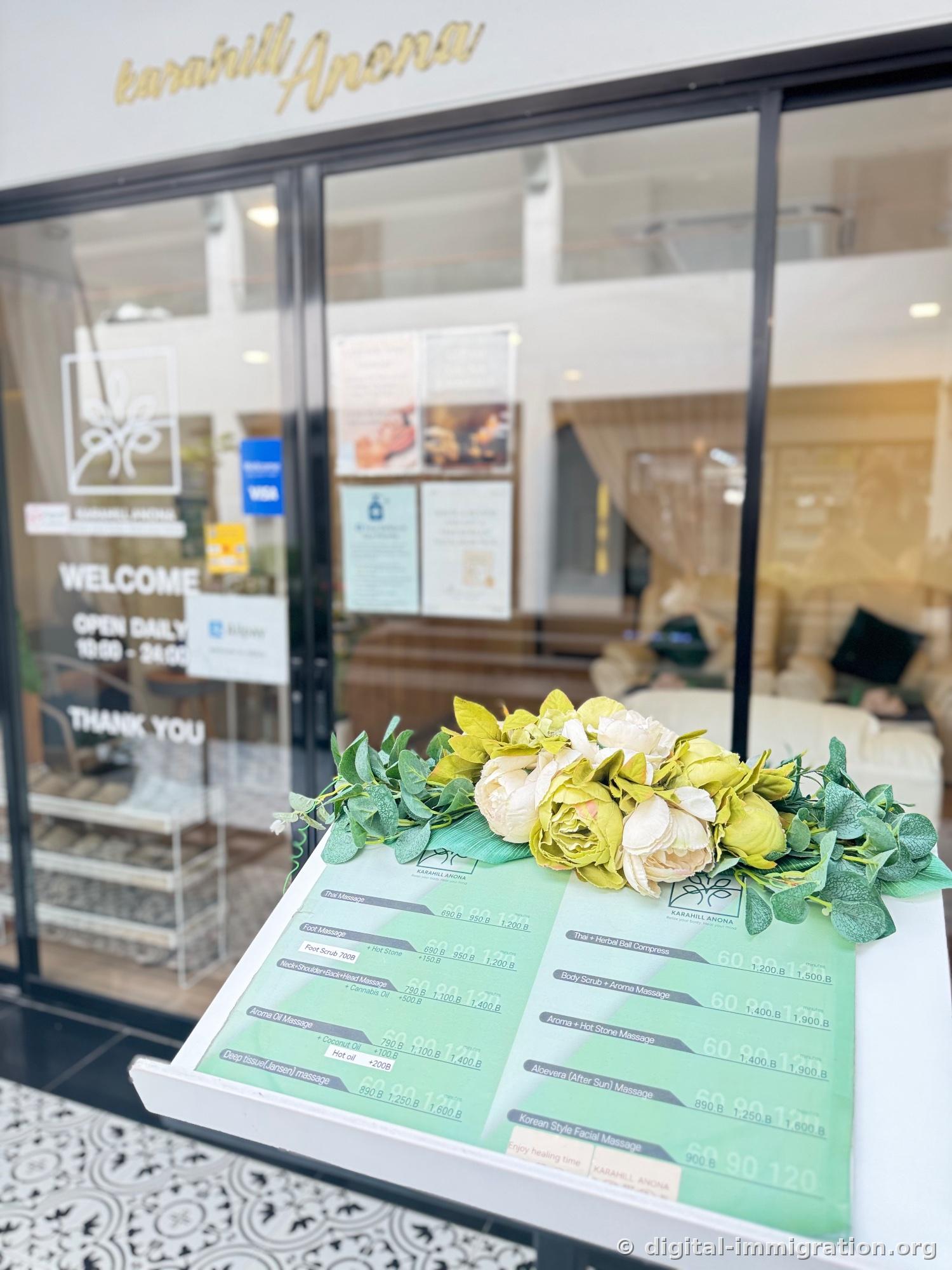 Green massage price list on a white stand next to the glass door with artificial decorations, karahill Anona gold sign and WELCOME notice visible in the background during daytime