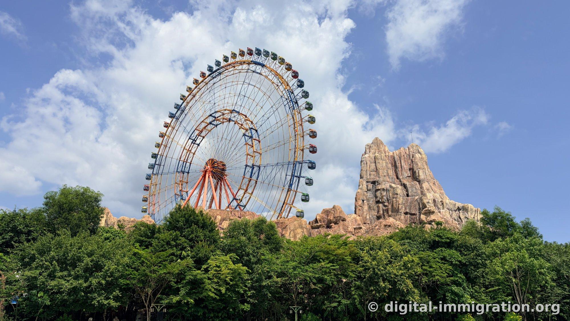 A colorful ferris wheel under blue sky with a red artificial rocky mountain beside it and dense trees in front