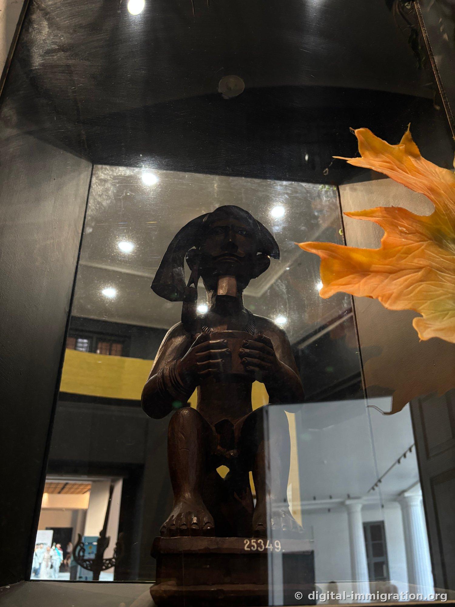 A wooden figure statue inside a glass display case kneeling and holding a bowl, with the museum courtyard and an orange artificial leaf reflected in the glass