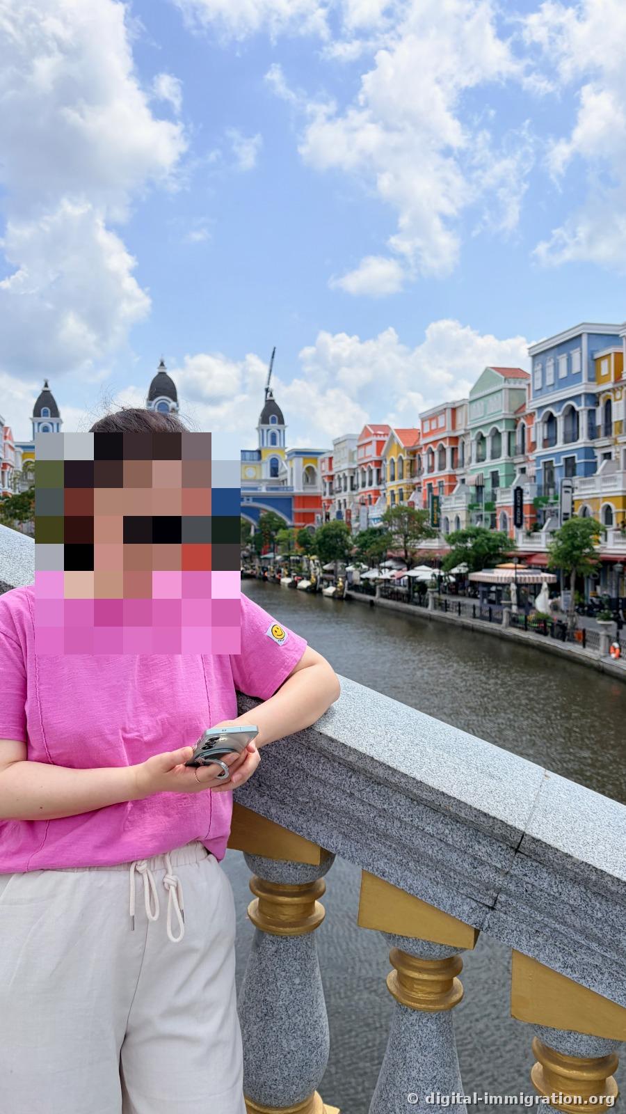 A person with arms resting on a bridge railing holding a phone, with shops and folded parasols lining the far canal bank