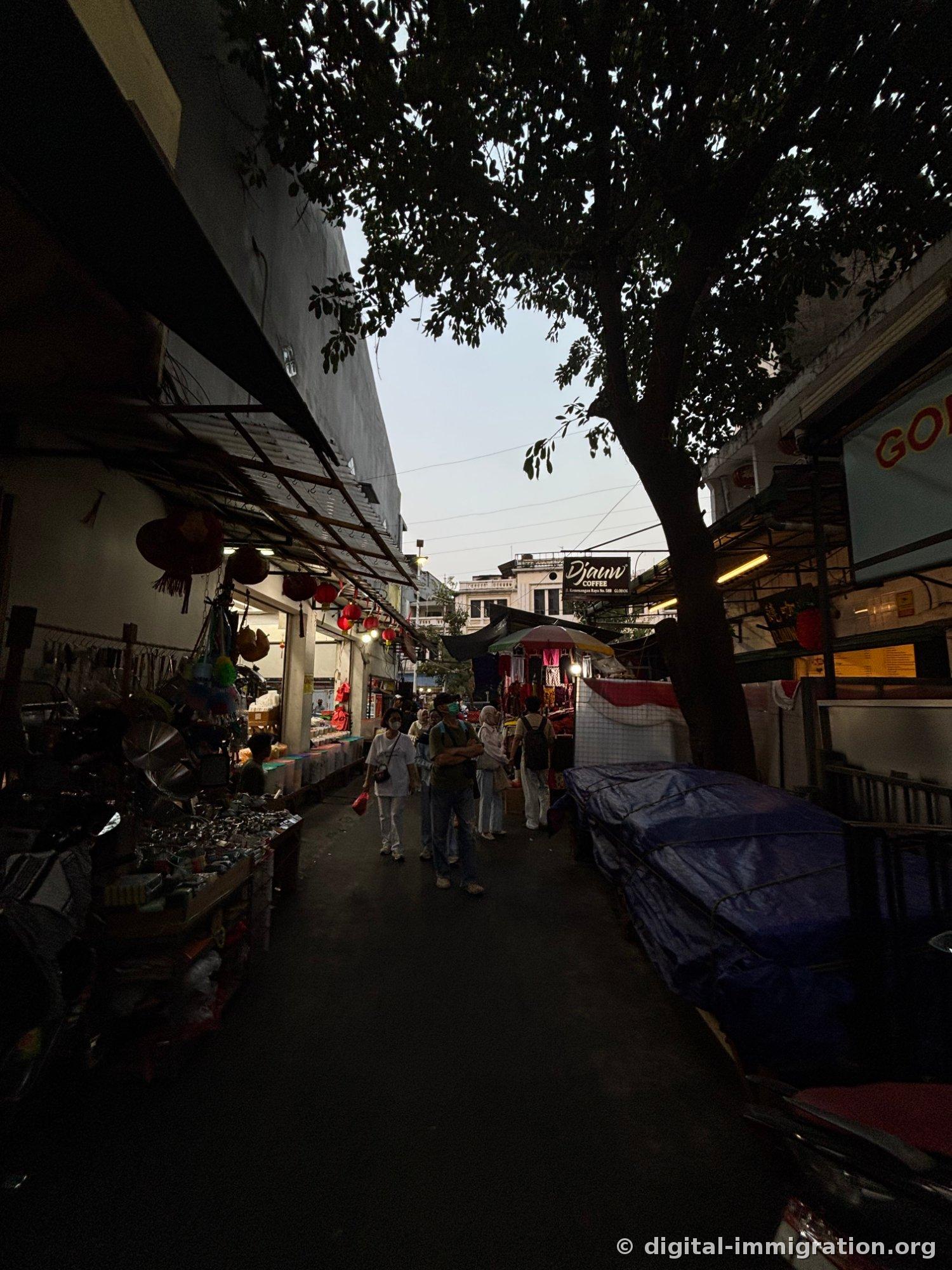 A narrow market alley at dusk, with pots and kitchen utensils hanging on the left stall and red lanterns draped low, with a 'Djauw Coffee' sign visible in the distance