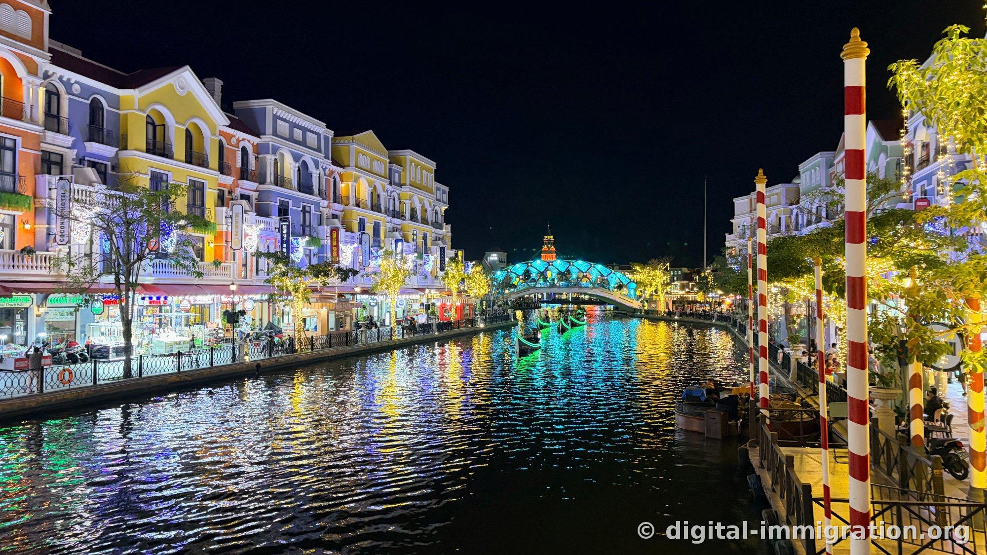 Night canal view with turquoise-lit arch bridge and an orange-lit clock tower in the distance, colorful buildings lit up on both sides