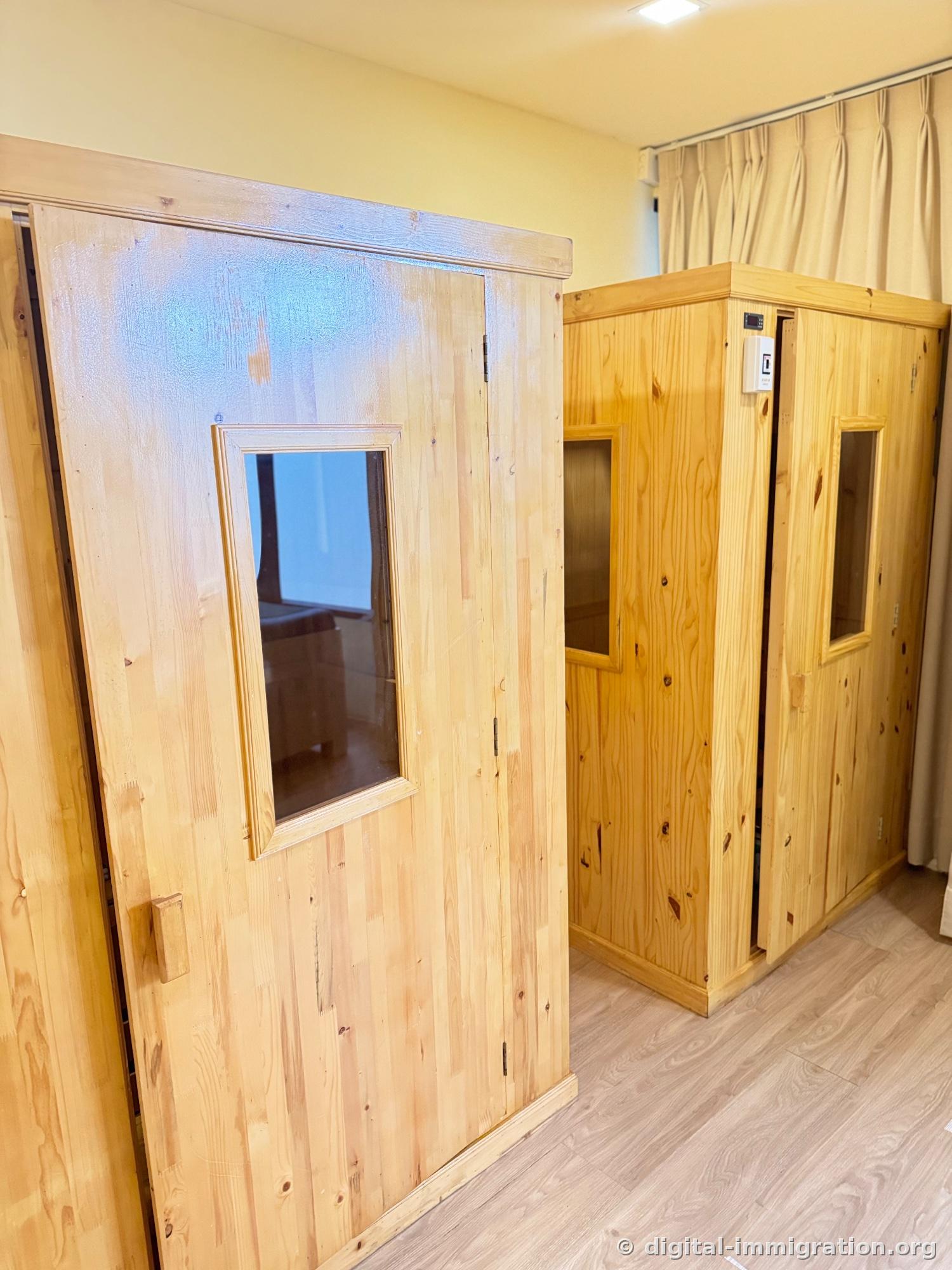 Two pine wood sauna booths installed side by side indoors, small glass window on the door and digital temperature controller visible