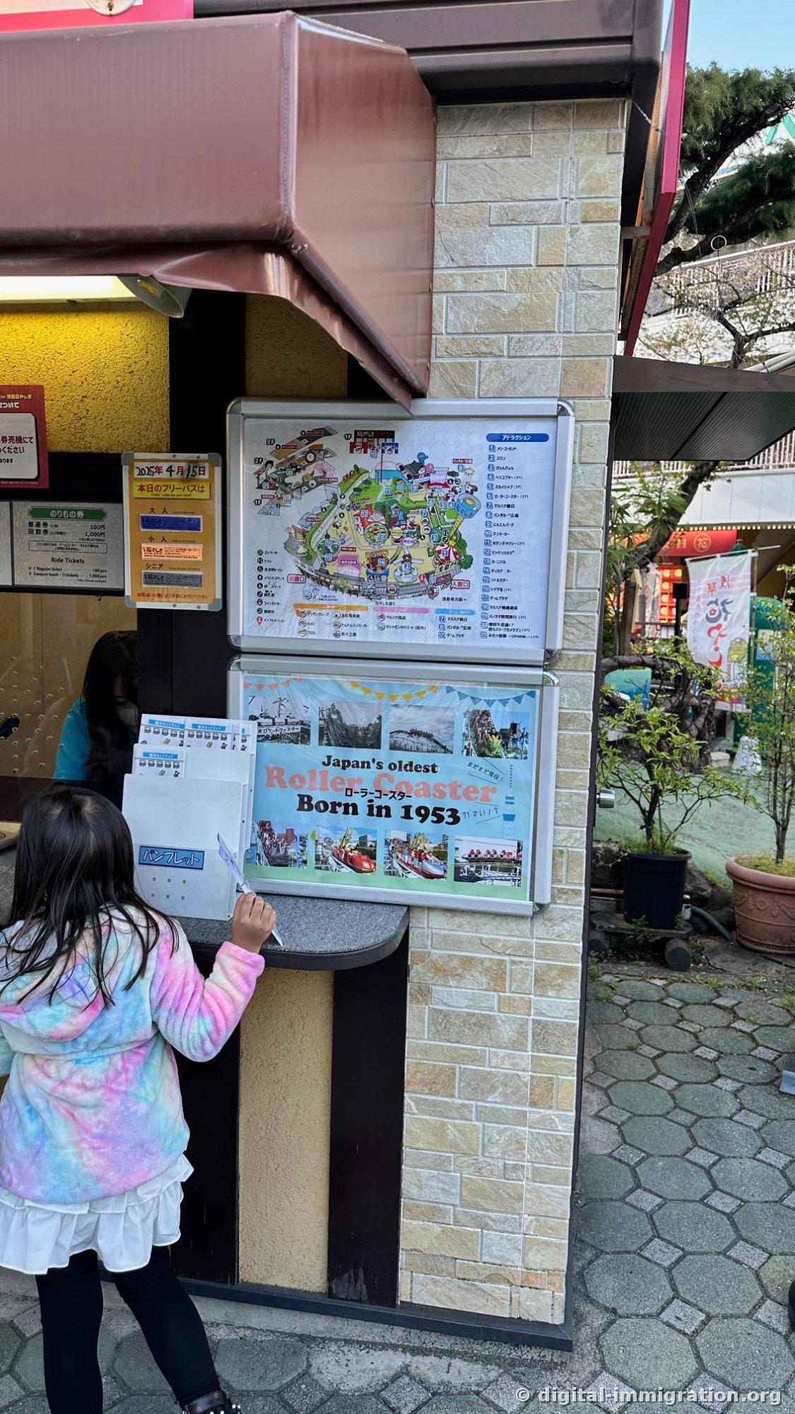 Park map and fee schedule posted on a pillar next to the Hanayashiki ticket window, with a 'Japan's oldest Roller Coaster Born in 1953' poster below, and a child pulling pamphlets in front