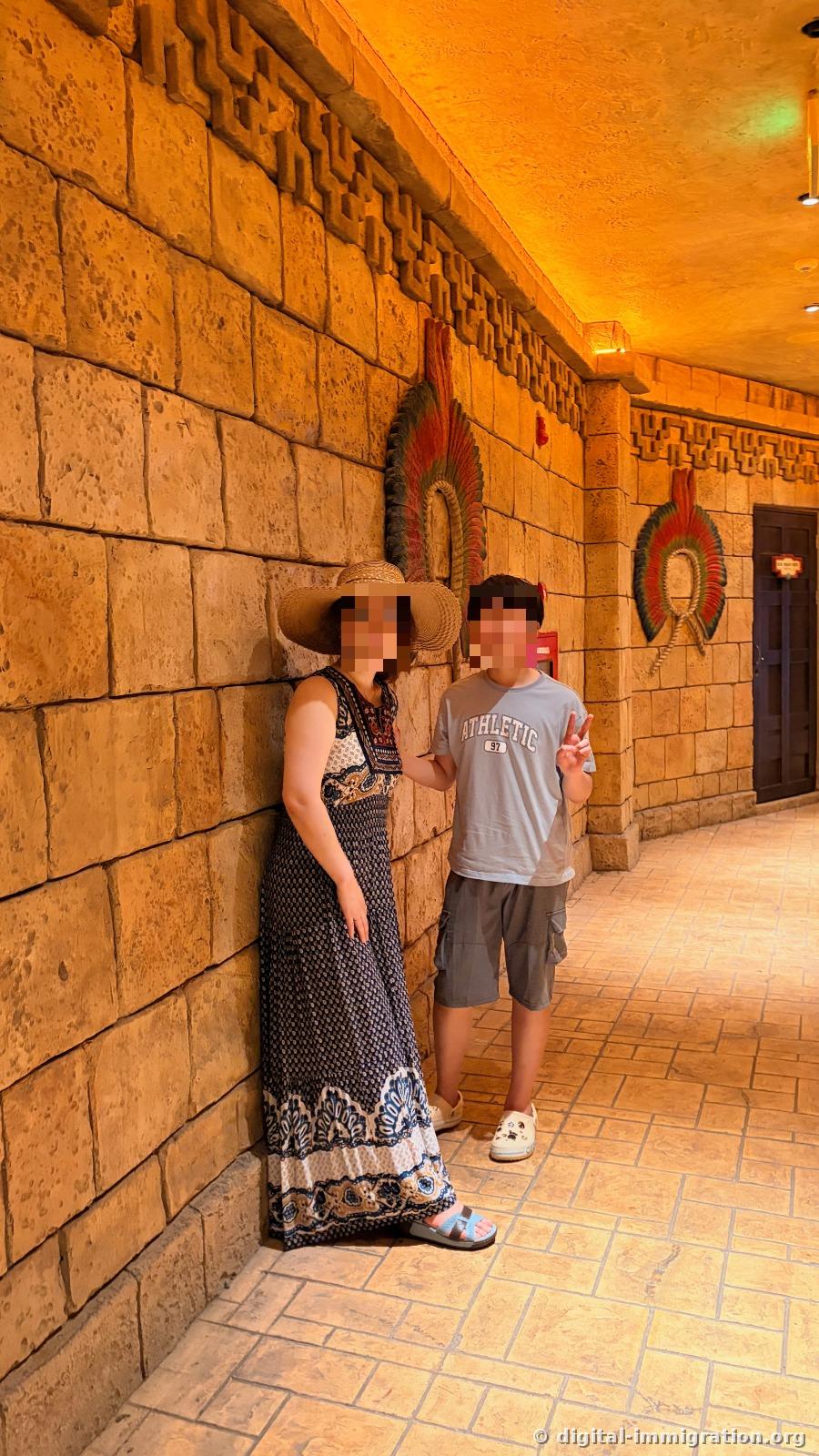 Two people in an indoor corridor with Mayan temple-style stone walls and feather decorations
