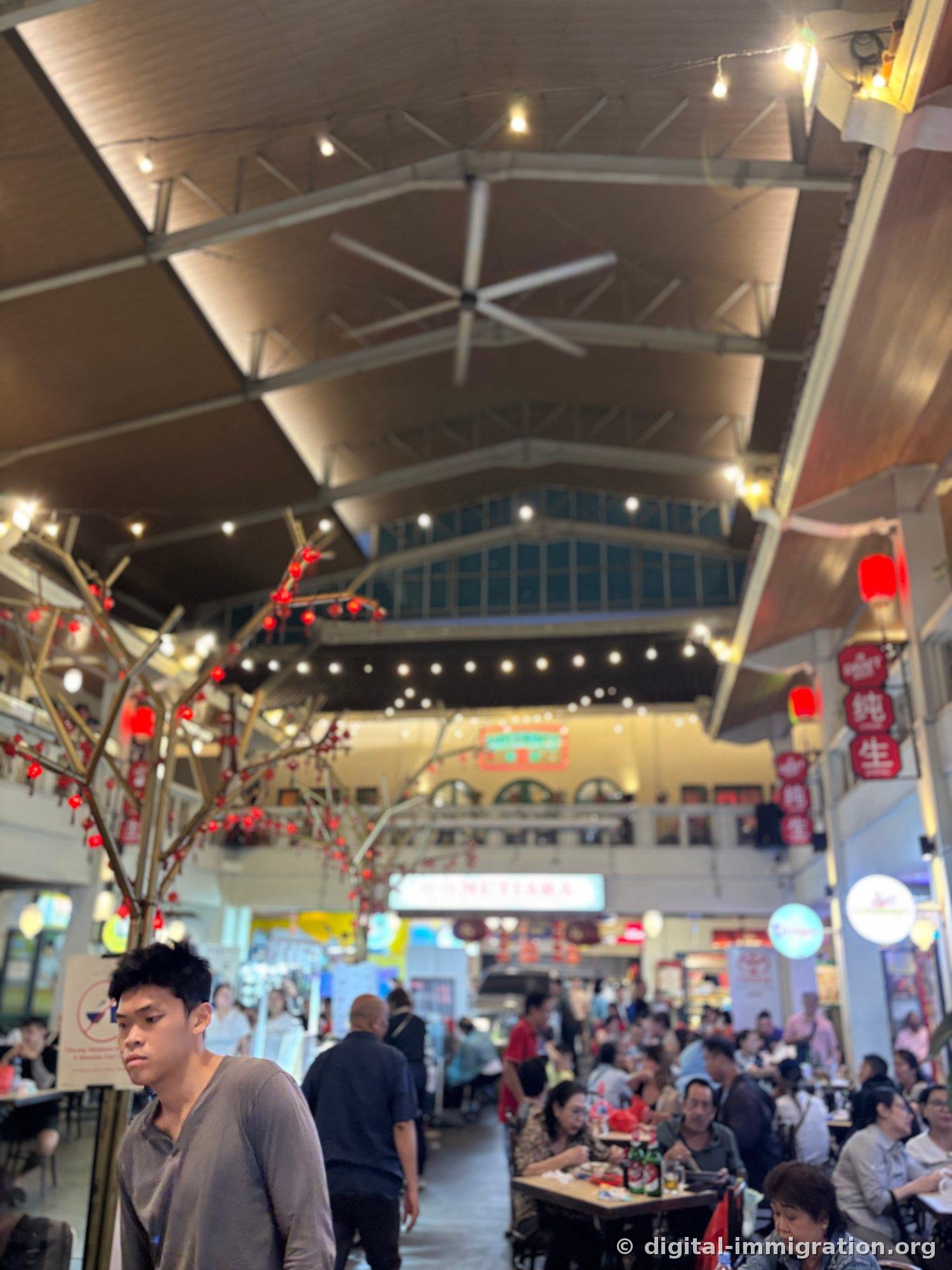 Interior of Petak Enam under high wooden ceiling and large ceiling fans, with red lanterns and hanging light bulbs, full dining tables, and an artificial tree with red berries