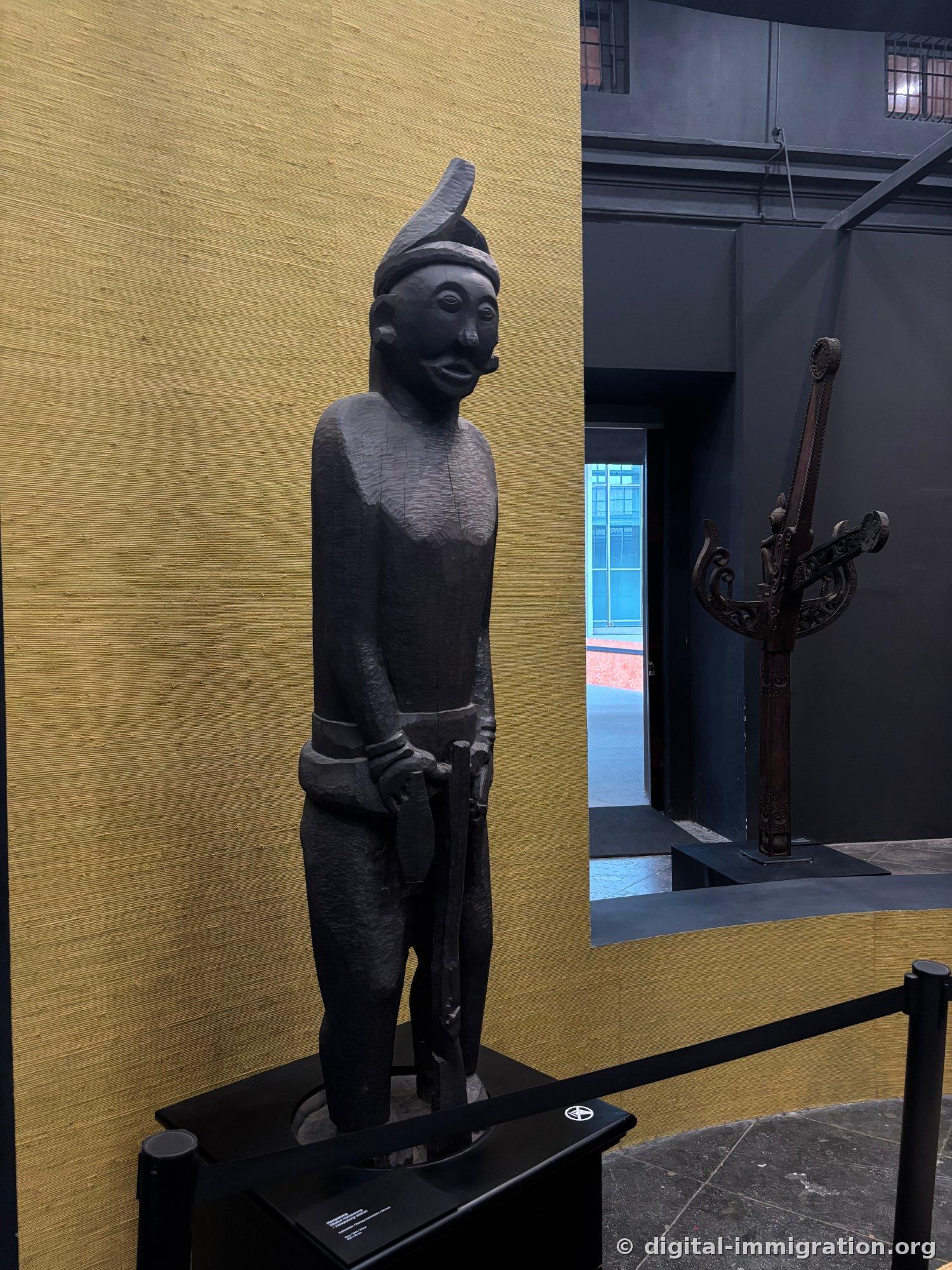 A black wooden ancestral male figure before the golden wall, wearing a pointed hat and leaning on a staff with both hands, with a carved red pillar visible behind