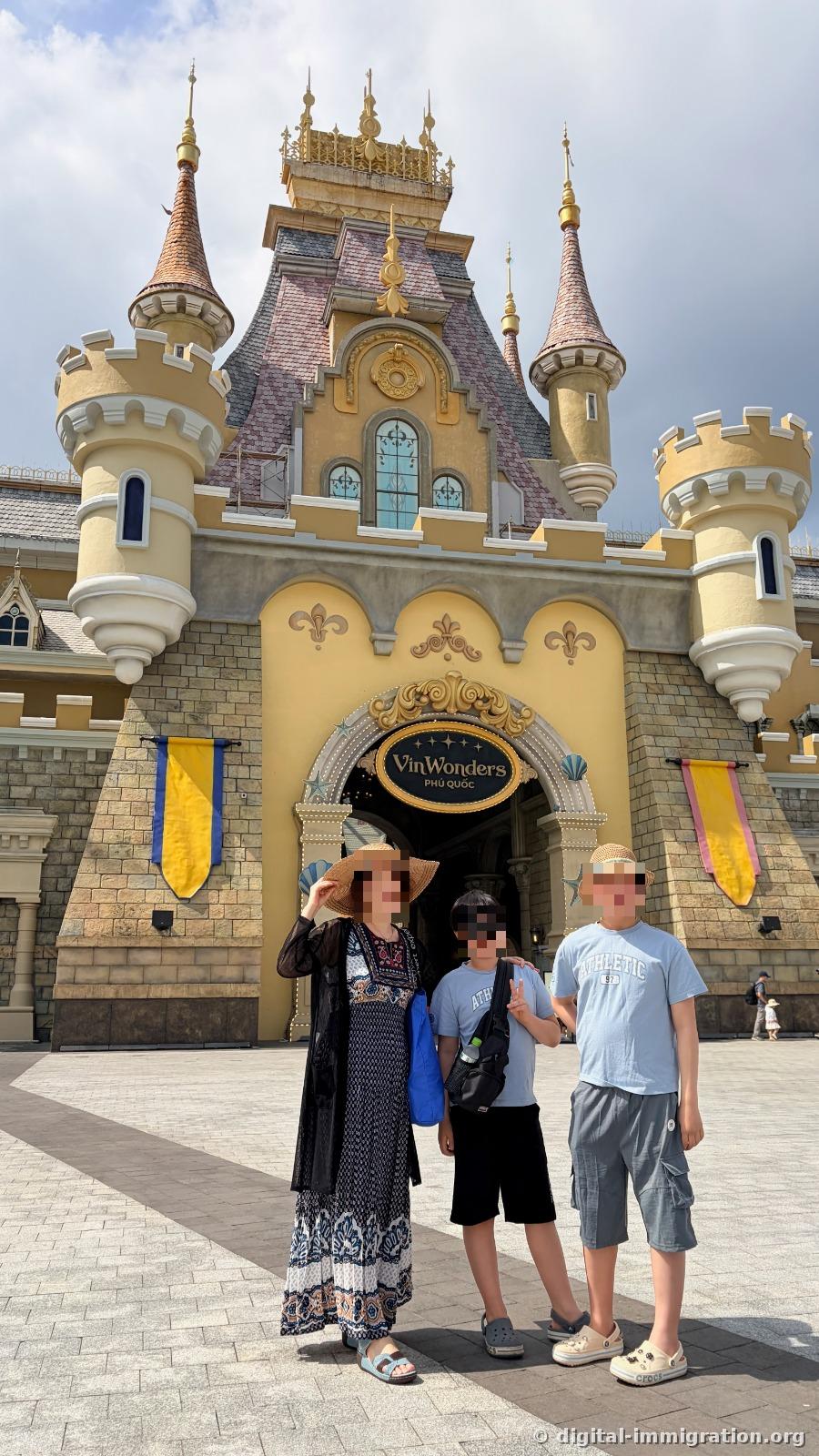 A family of three standing in front of the yellow castle-shaped main gate with the 'VinWonders PHÚ QUỐC' sign