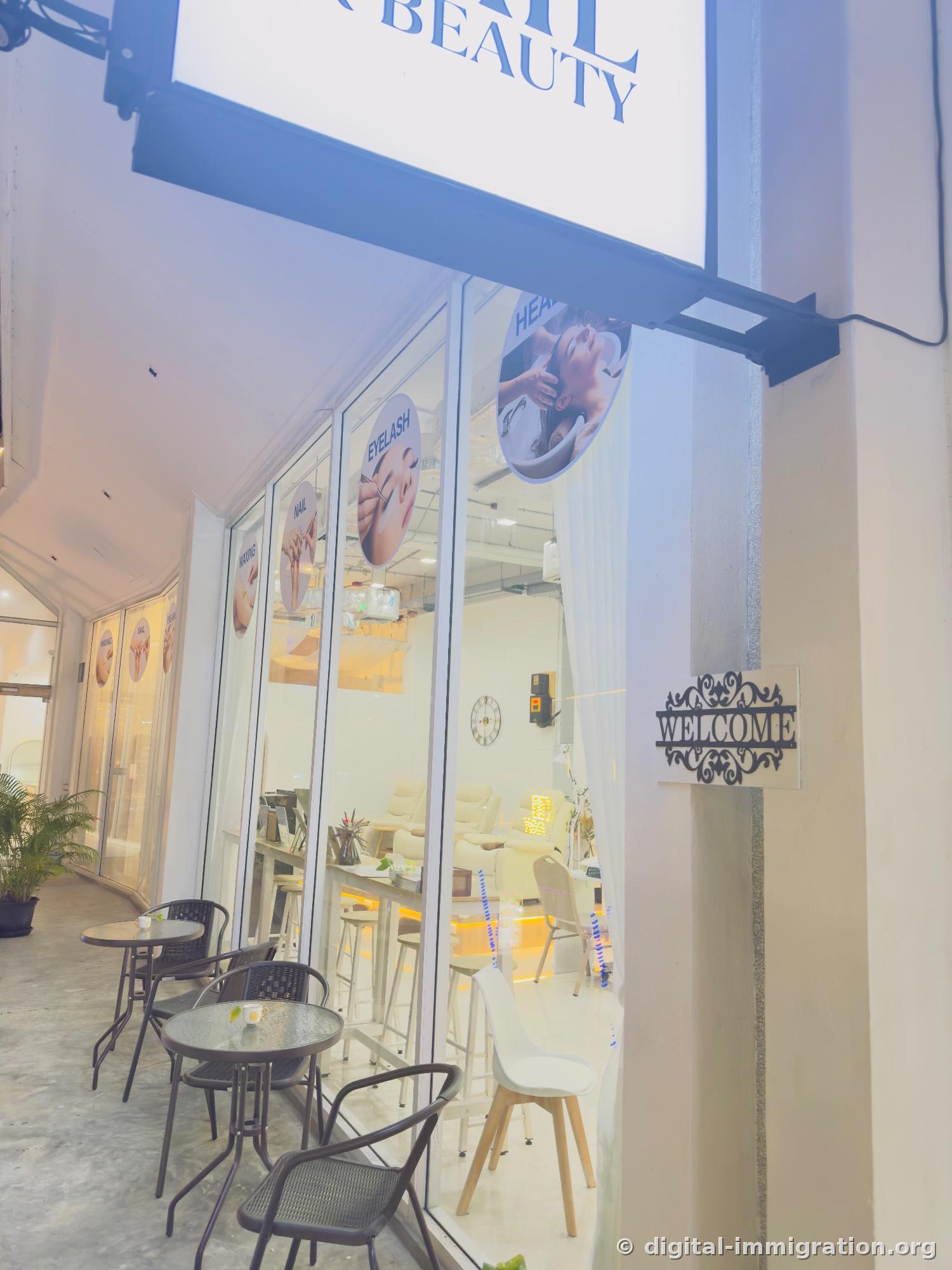Evening storefront exterior with NAIL & BEAUTY illuminated sign and circular photo icons for WAXING·NAIL·EYELASH·HEAD SPA on glass window, metal WELCOME sign on pillar and outdoor tables