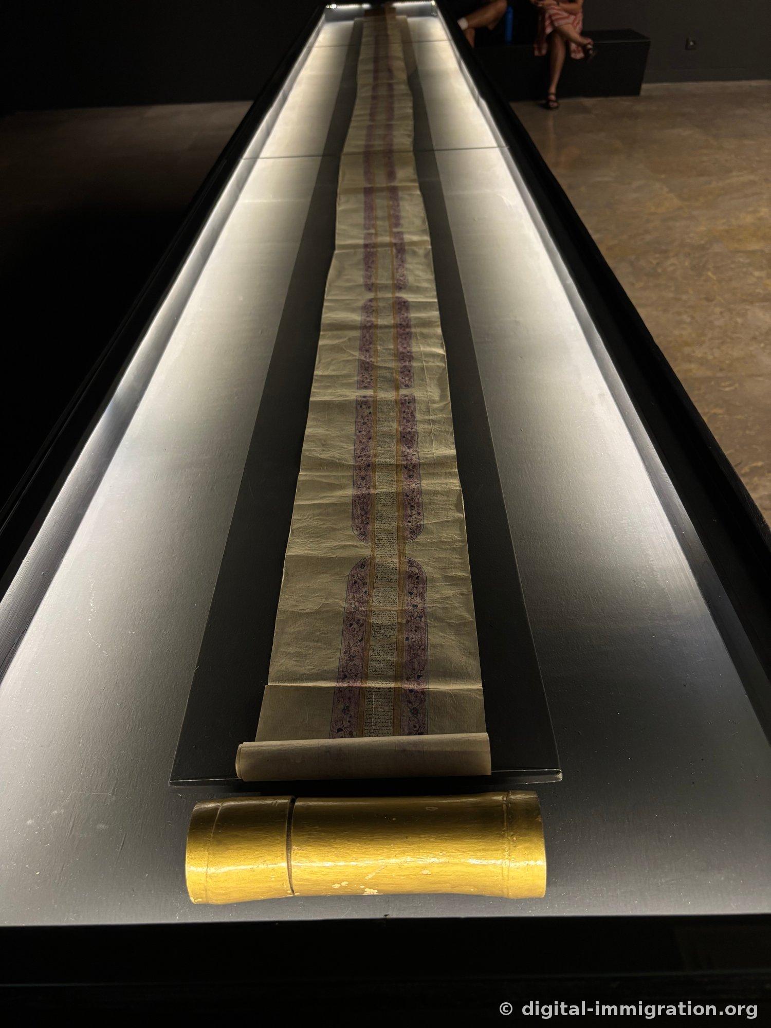 A long glass display case with an unrolled yellowish scroll manuscript inside, featuring purple border decoration and dense handwritten text, with a yellow storage tube in front