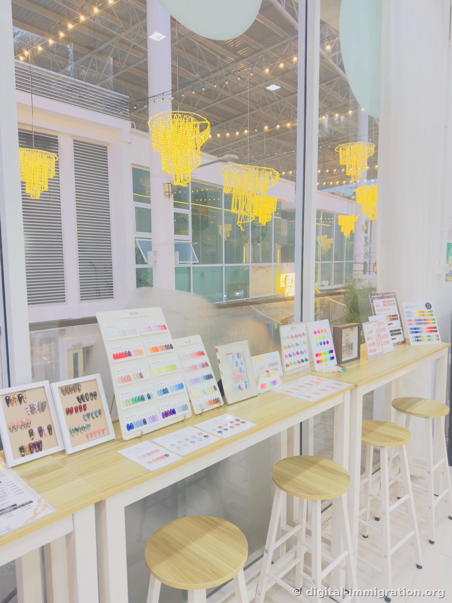 Several nail color charts displayed on natural wood bar table by the window, framed art design samples, wooden stools, yellow chandelier lighting beyond the glass window