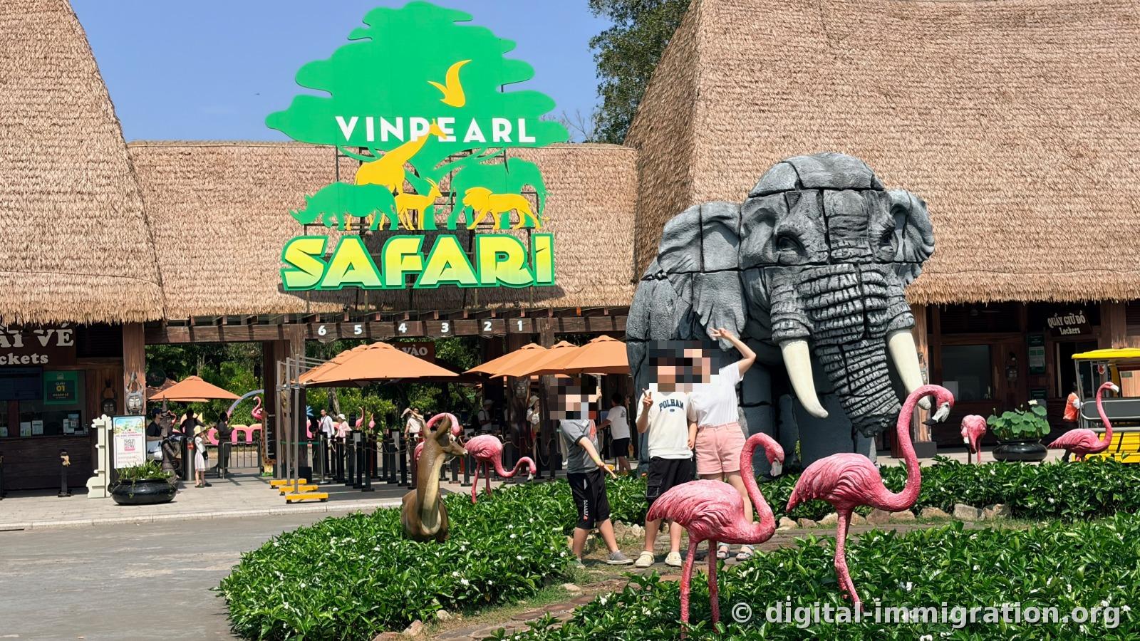 Green VINPEARL SAFARI sign hanging above the straw-roofed ticket office, with gray elephant and pink flamingo sculptures in front where children pose at the main entrance