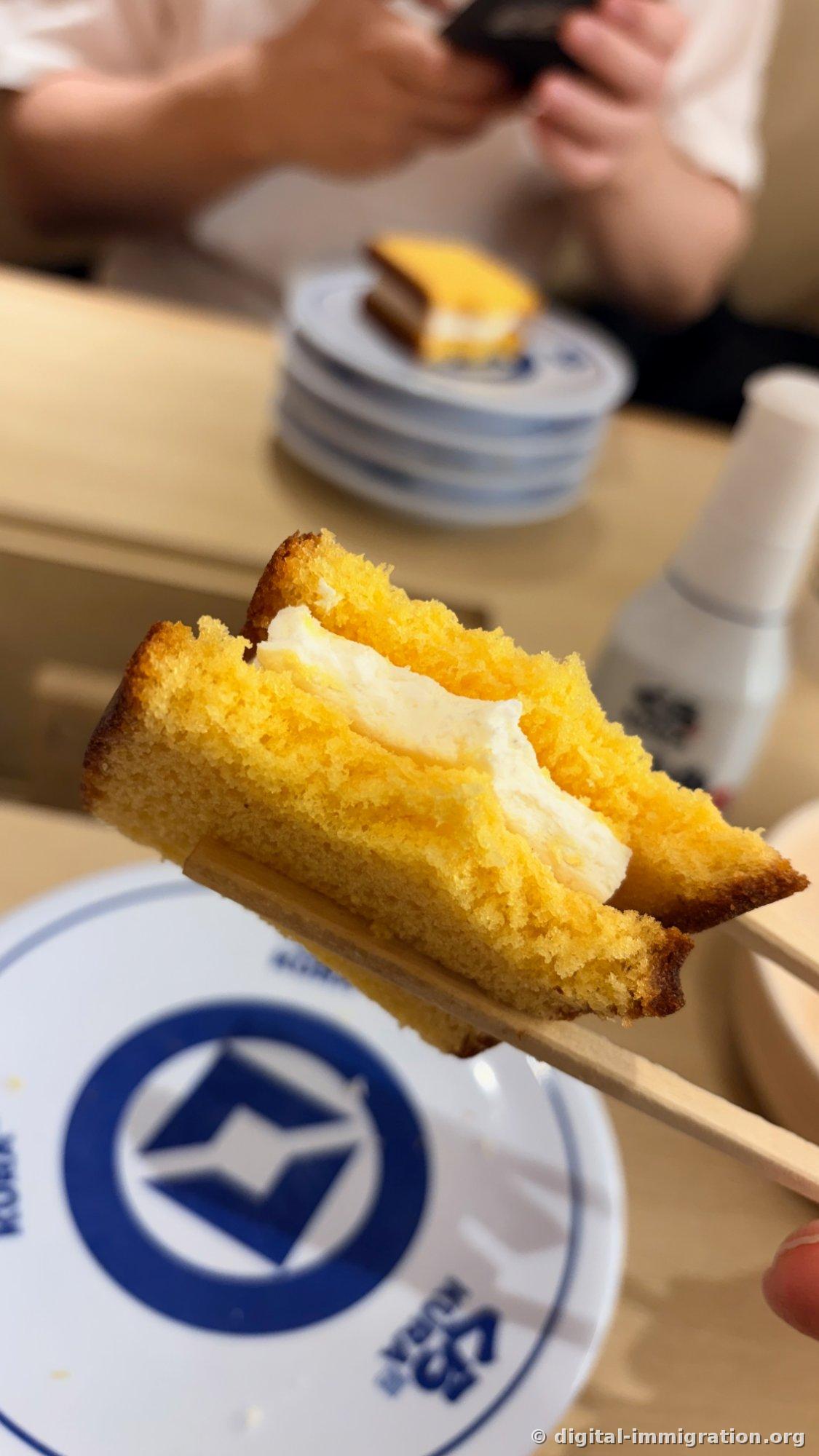 Gâteau Castella avec une bouchée prise, soulevé avec des baguettes. La crème s'écoule sur les côtés et la tour d'assiettes et un autre gâteau sont visibles à l'arrière
