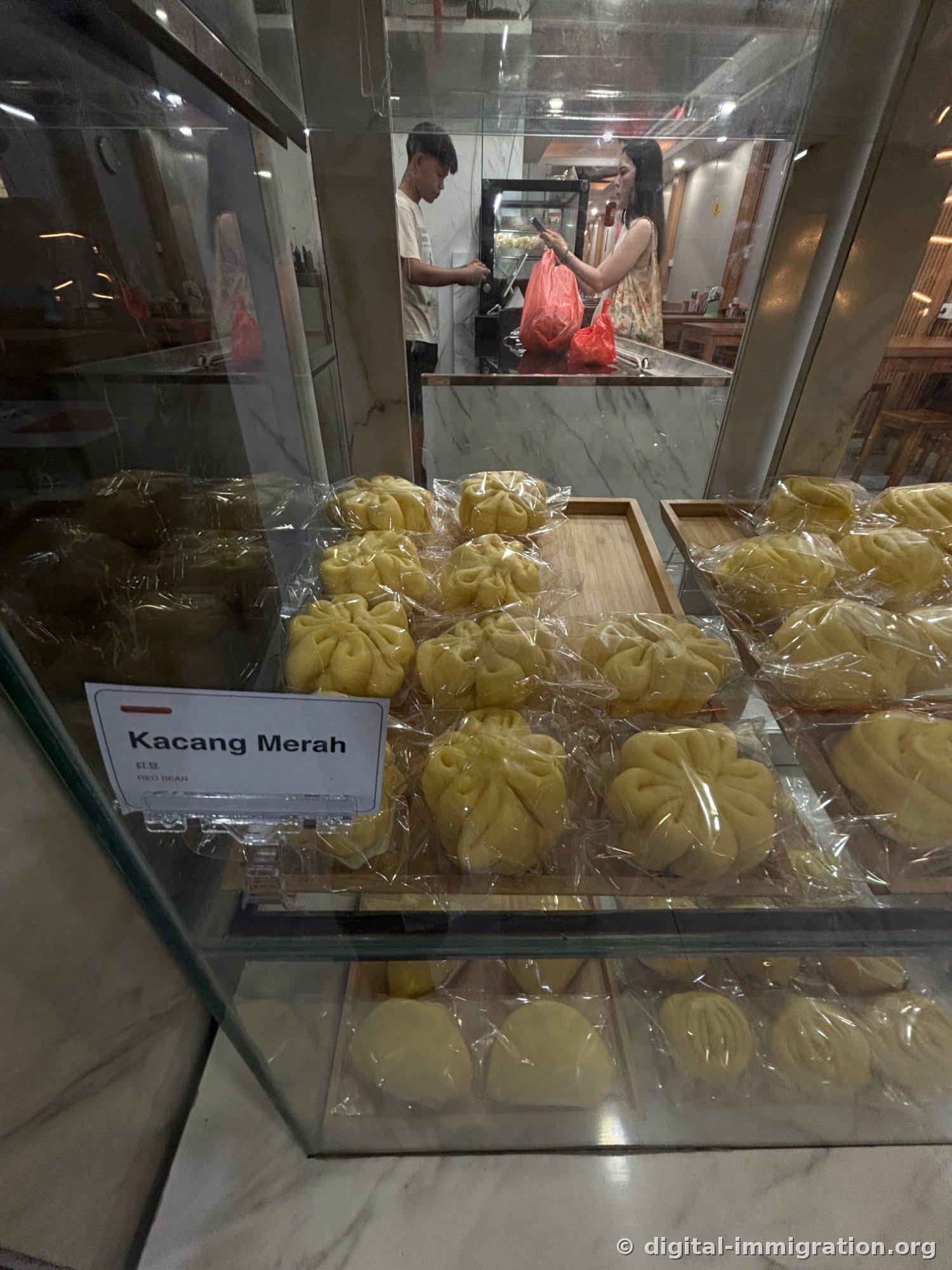 À l'intérieur d'une vitrine en verre, des baozi jaunes emballés en plastique sont arrangés par type, avec une étiquette inscrite Kacang Merah RED BEAN devant