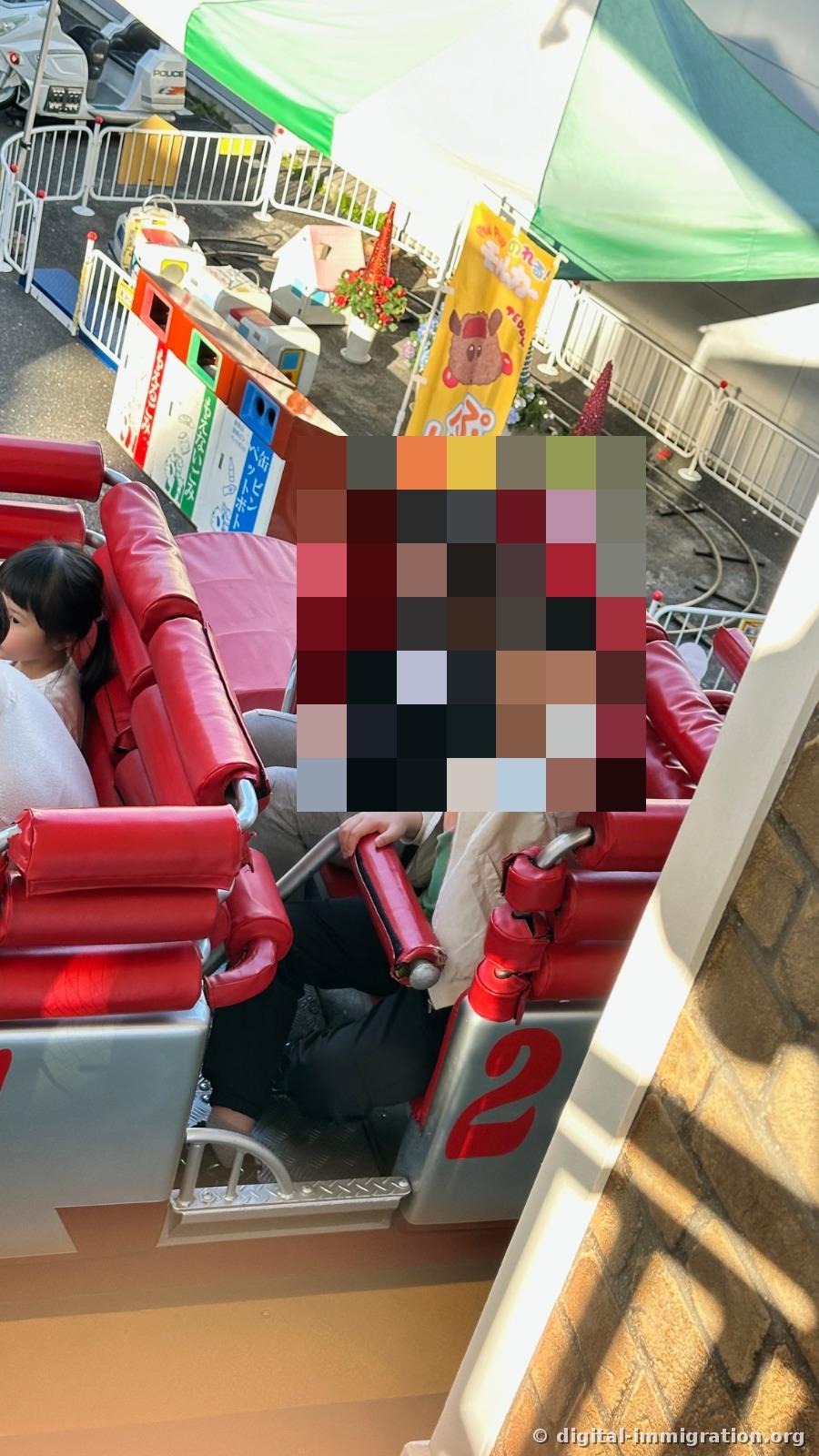 Passengers in red seat 2 of the roller coaster, and beyond the fence: recycling bins, tent, police car