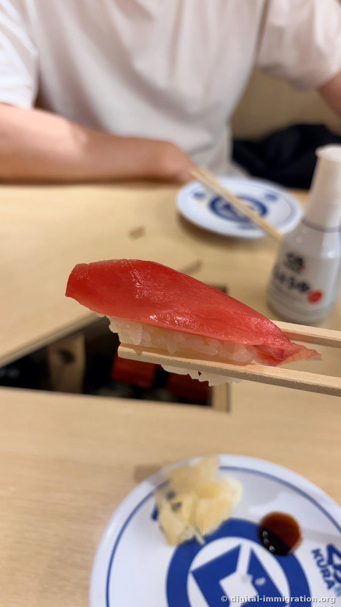 Sushi au thon soulevé avec des baguettes à la place du comptoir. À l'arrière, le logo Kura Sushi et la bouteille de sauce soja blanche avec しょうゆ écrit dessus et l'ouverture du rail
