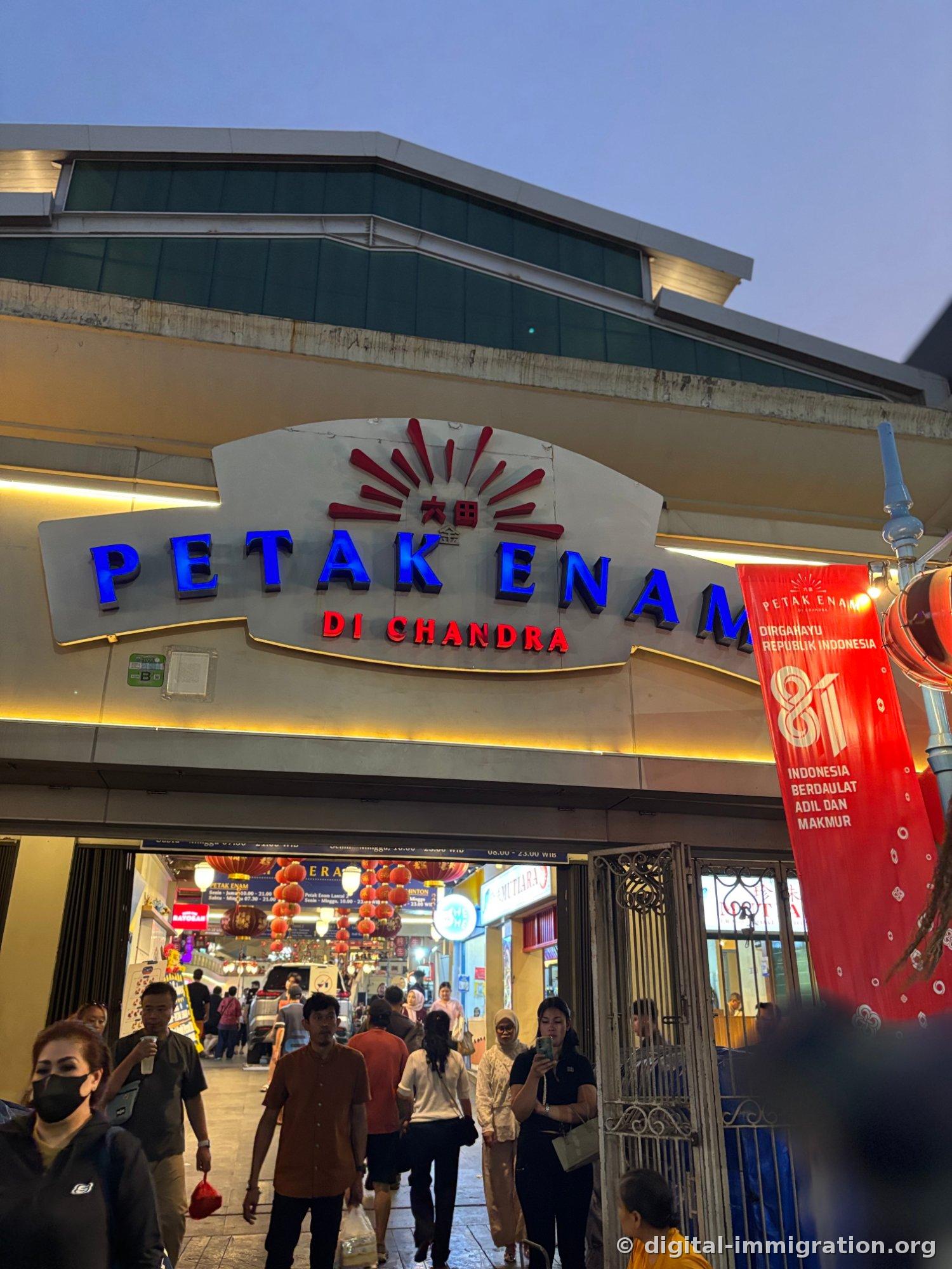 Building entrance with blue neon 'PETAK ENAM DI CHANDRA' signage, red Independence Day banner, hallway with lanterns inside, and people coming and going