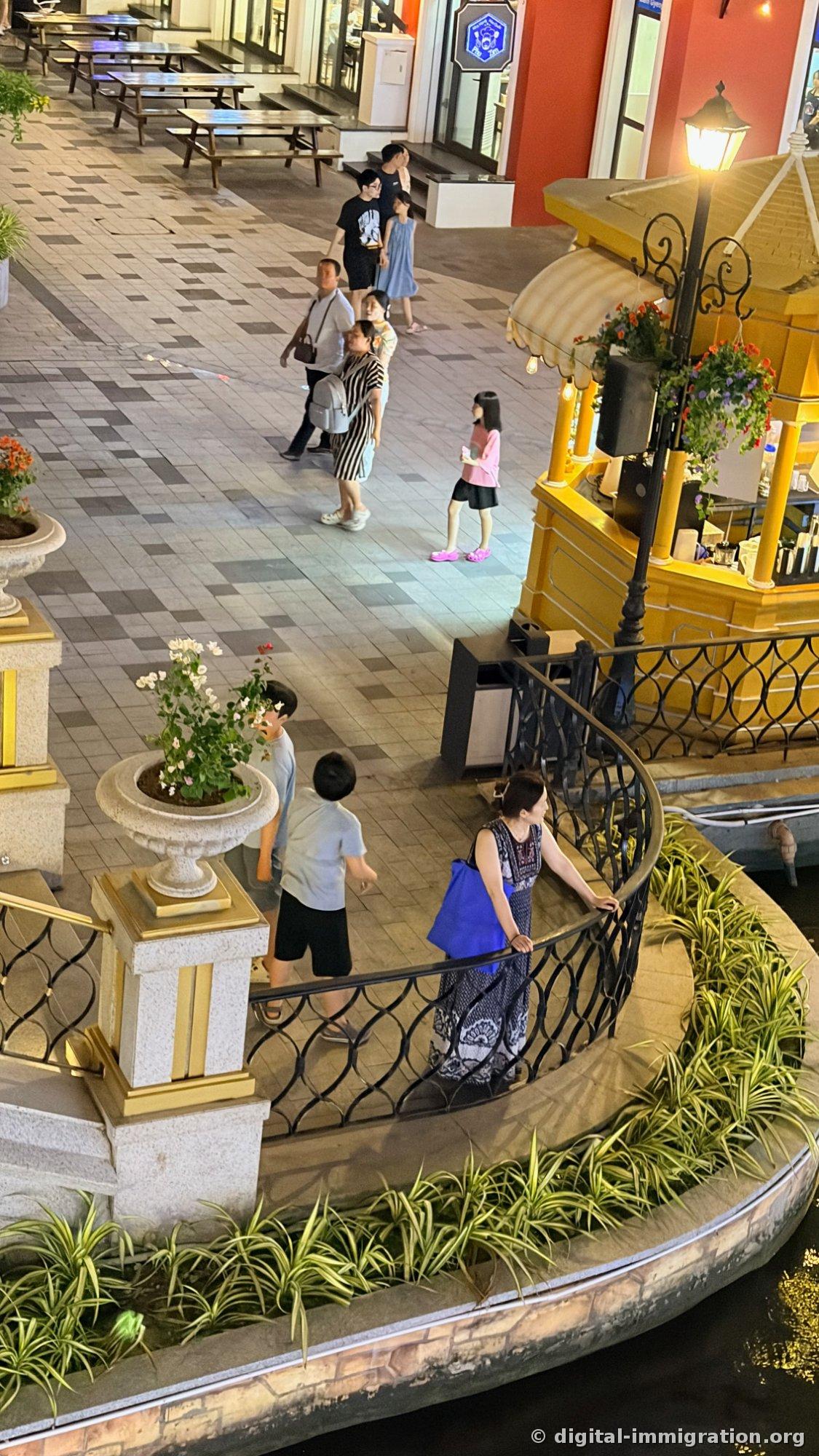 Looking down at the night promenade from above with a yellow wooden kiosk, street lamps, and people using the curved stairs