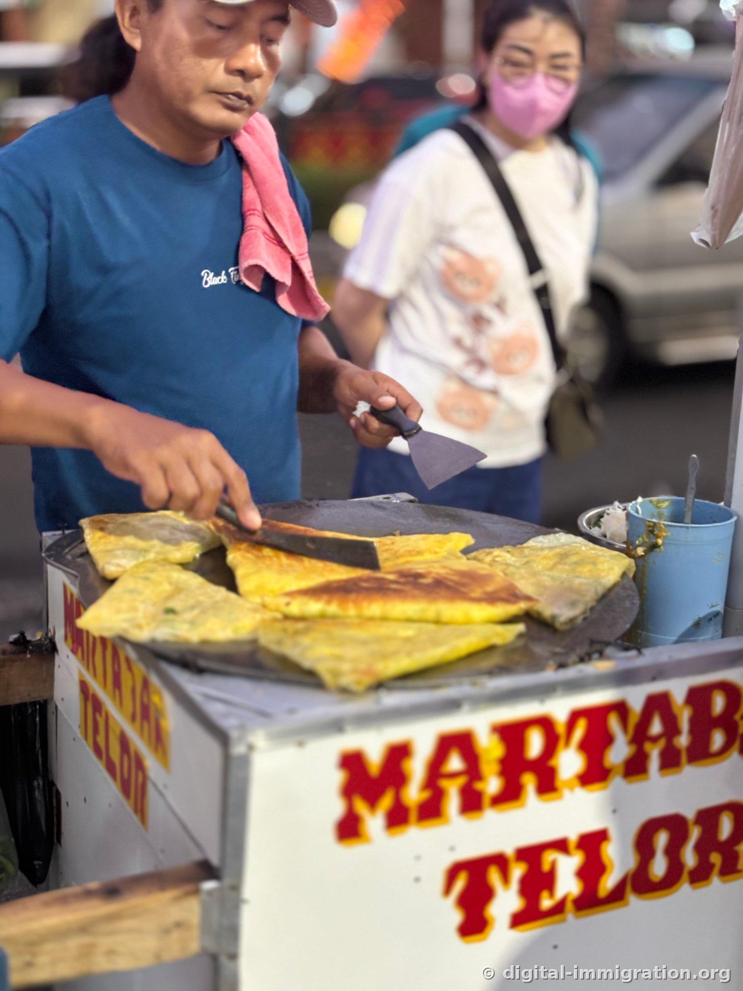 A vendor in a blue t-shirt with a pink towel draped over his shoulder frying Martabak Telor on the griddle of a 'MARTABAK TELOR' hand-cart using two spatulas, with a customer waiting behind