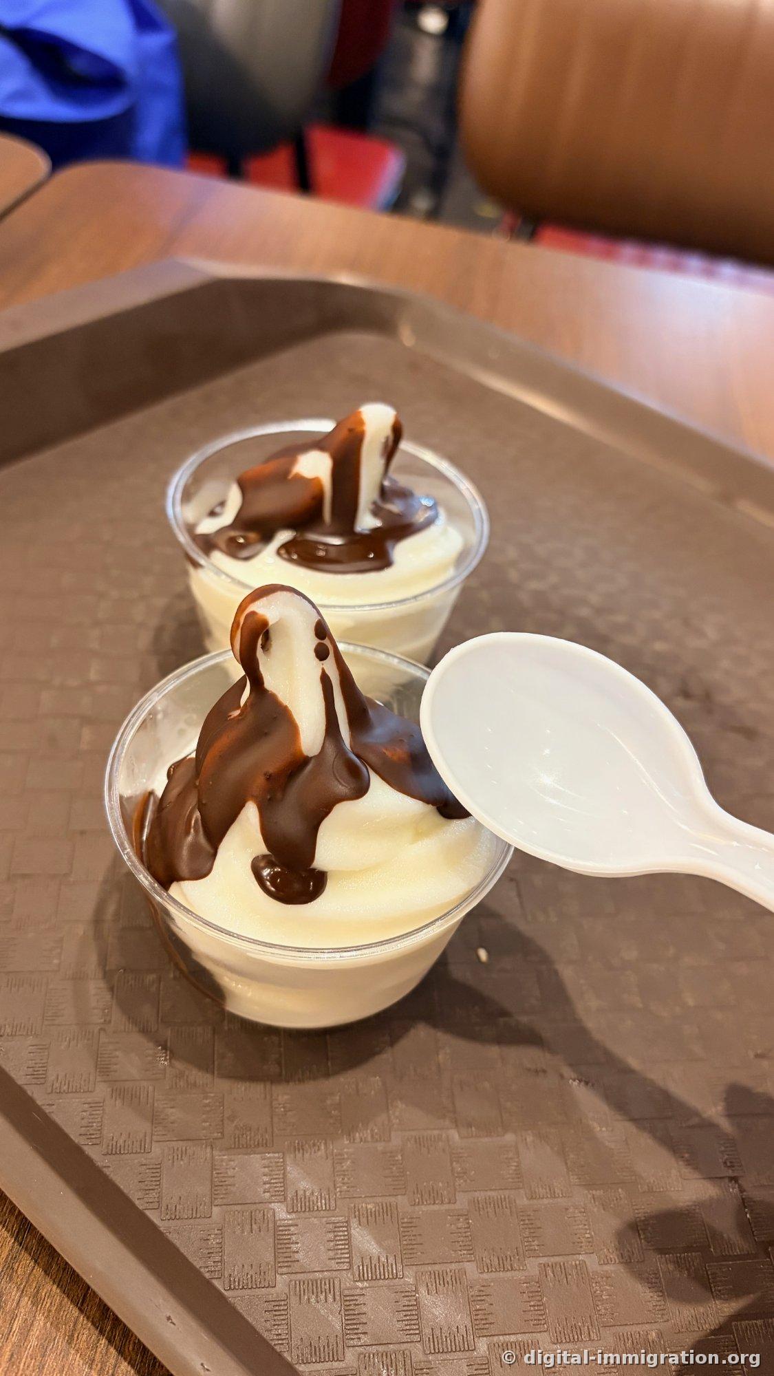 Two cups of soft serve ice cream topped with chocolate sauce and white plastic spoons on a tray