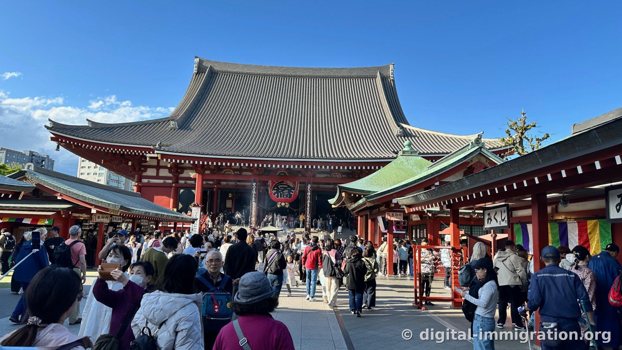 The front of the Senso-ji main hall with a red lantern bearing the character '福', stamp and fortune-telling booths on both sides, and a courtyard full of visitors
