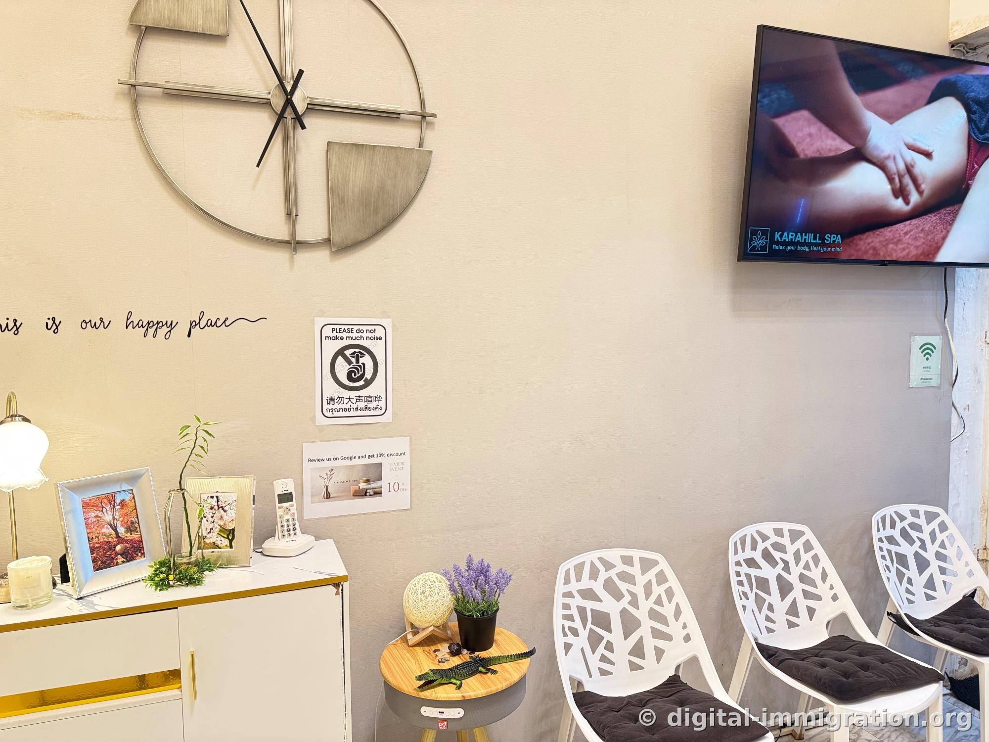 Metal wall clock and 'this is our happy place' lettering, multilingual notice requesting quiet, Google review 10% discount sign, wall-mounted TV and three white plastic chairs in waiting area
