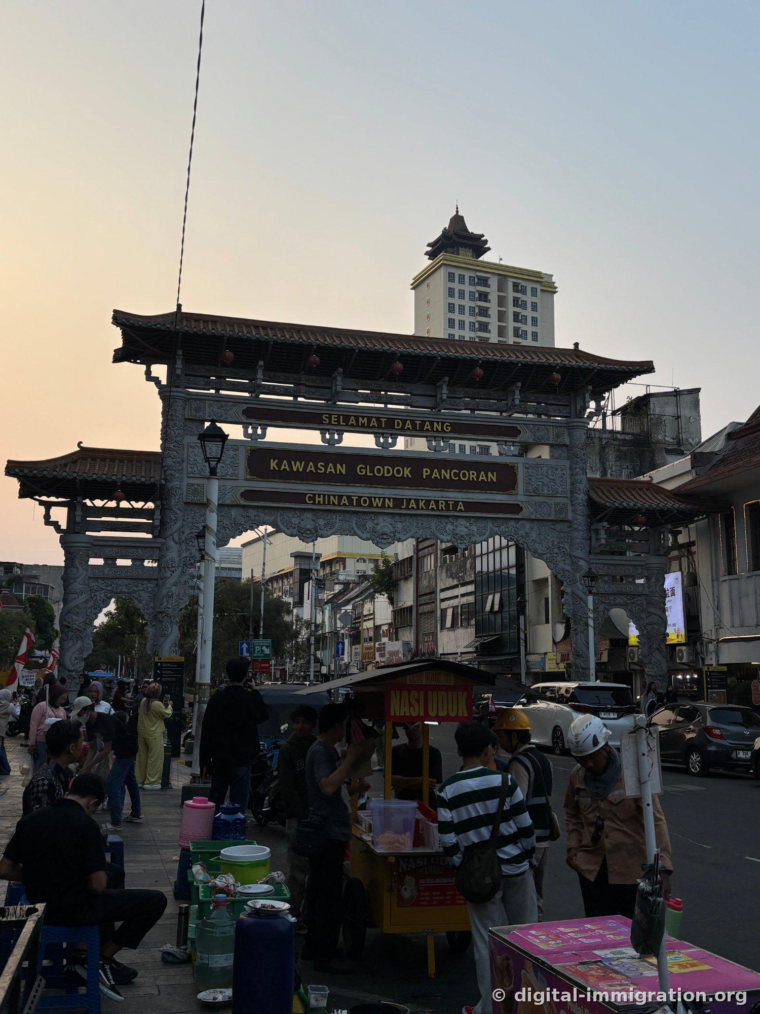 A gray stone archway with 'SELAMAT DATANG KAWASAN GLODOK PANCORAN CHINATOWN JAKARTA' inscribed on it at sunset, with a Nasi Uduk food cart below, and a high-rise apartment with Chinese-style roof eaves visible in the background