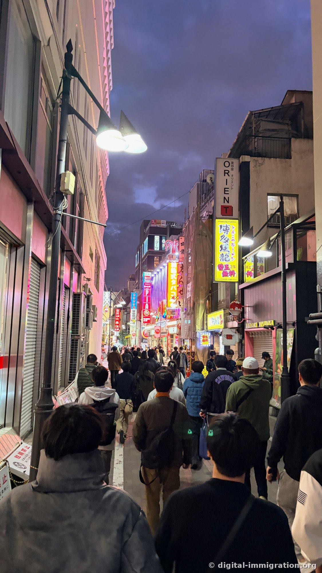 Une foule remplissant une ruelle étroite avec des enseignes au néon alignées de chaque côté, l'enseigne du salon de beauté « ORIENT » et une enseigne de ramen jaune Hakata visibles à droite