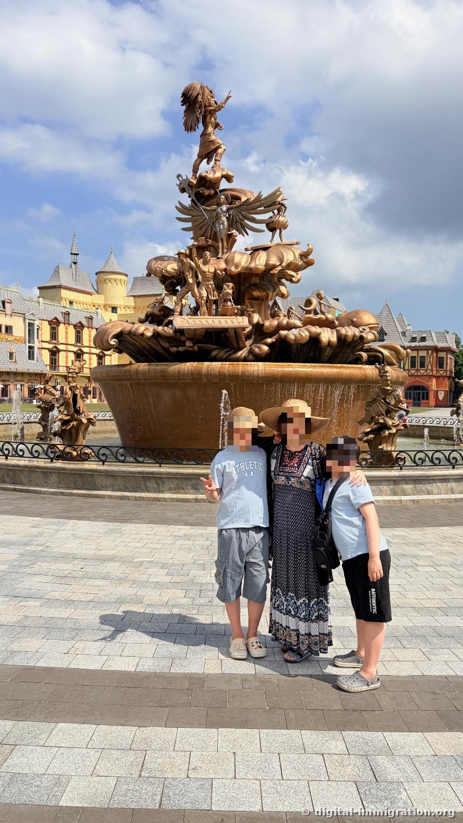 Three people standing in front of a large golden fountain sculpture with layered figures, with European-style buildings in the background