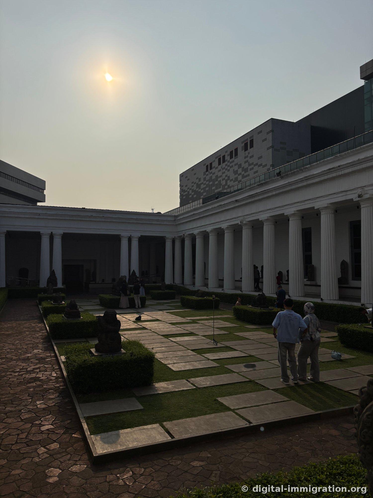 The museum courtyard surrounded by a white-columned colonnade, with grass and stone in a checkered pattern and visitors standing