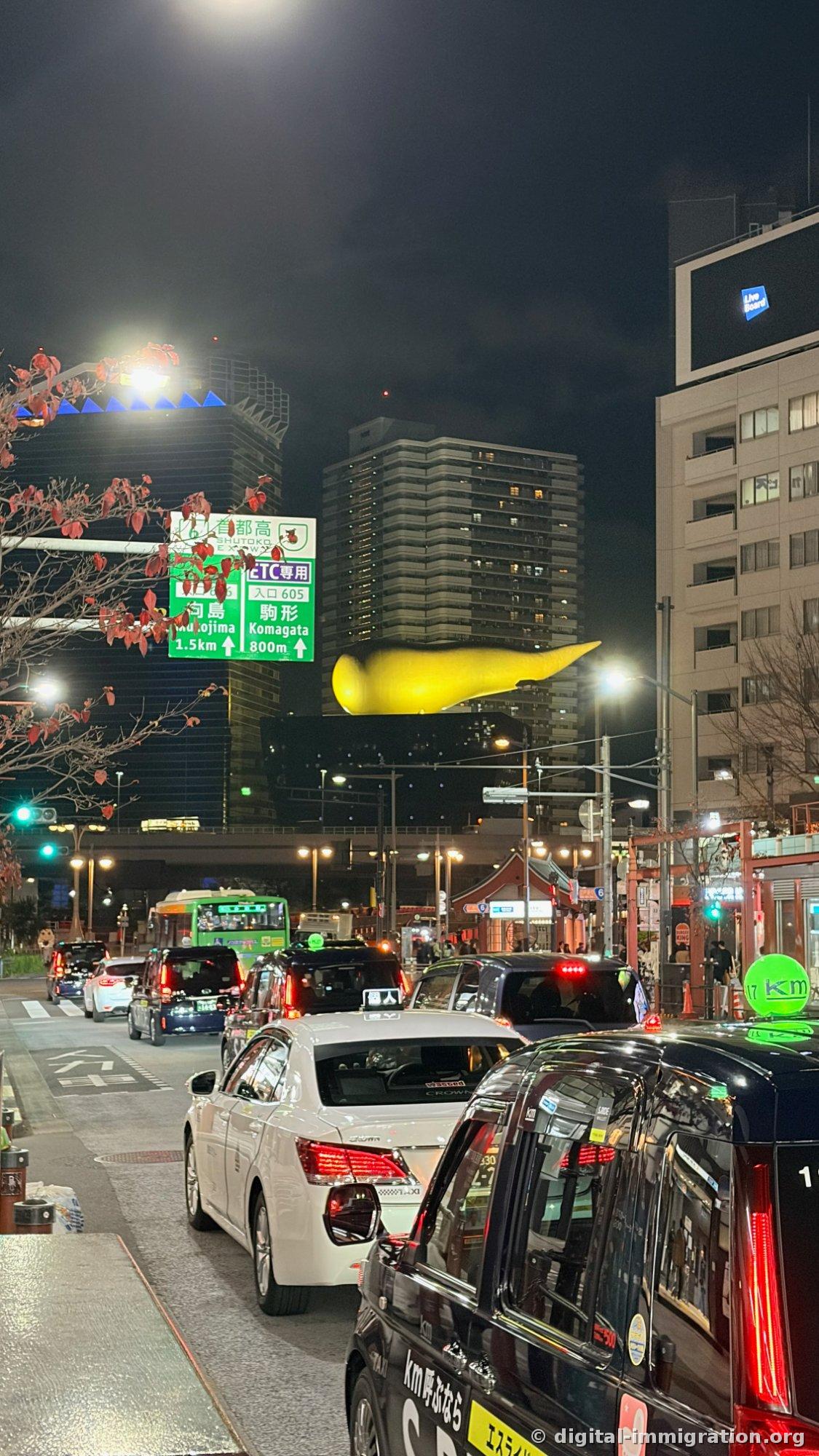 Azumabashi Intersection at night. Black and white taxis line up, with the golden flame sculpture lit in yellow behind them and a capital expressway sign visible.