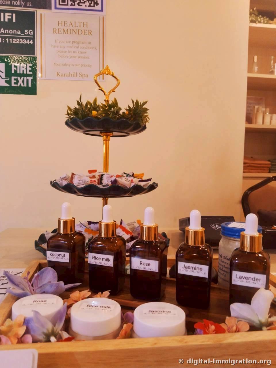 Five brown oil bottles labeled coconut, Rice milk, Rose, Jasmine, Lavender on a wooden tray with matching scrub containers, two-tiered tray of milk candies and HEALTH REMINDER notice in the background
