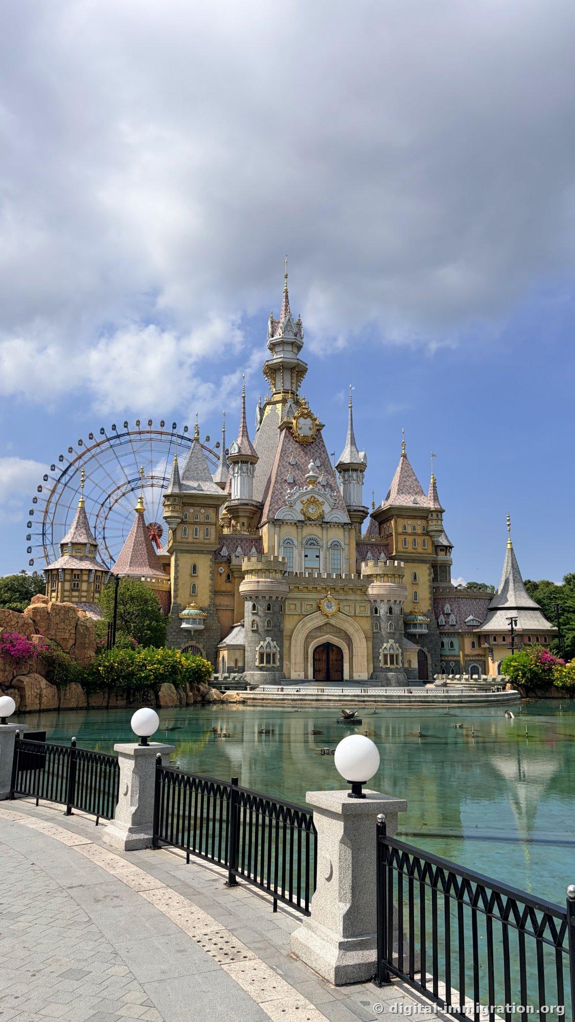 A European-style castle viewed across a lake in daylight with a large ferris wheel behind