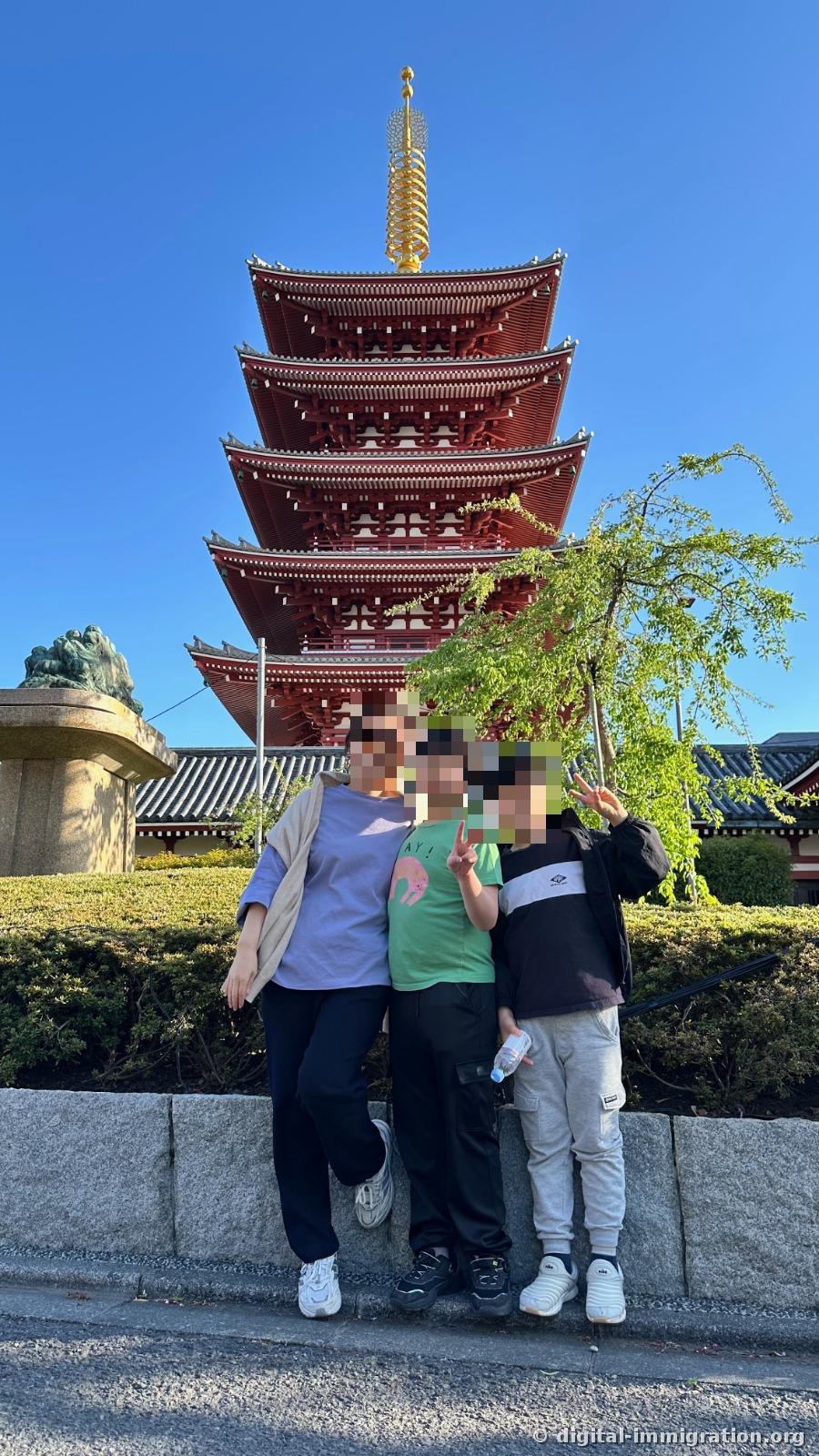 Three people standing in front of the five-story pagoda at the low stone wall, with a lion statue pedestal on the left and a tree with new leaves on the right
