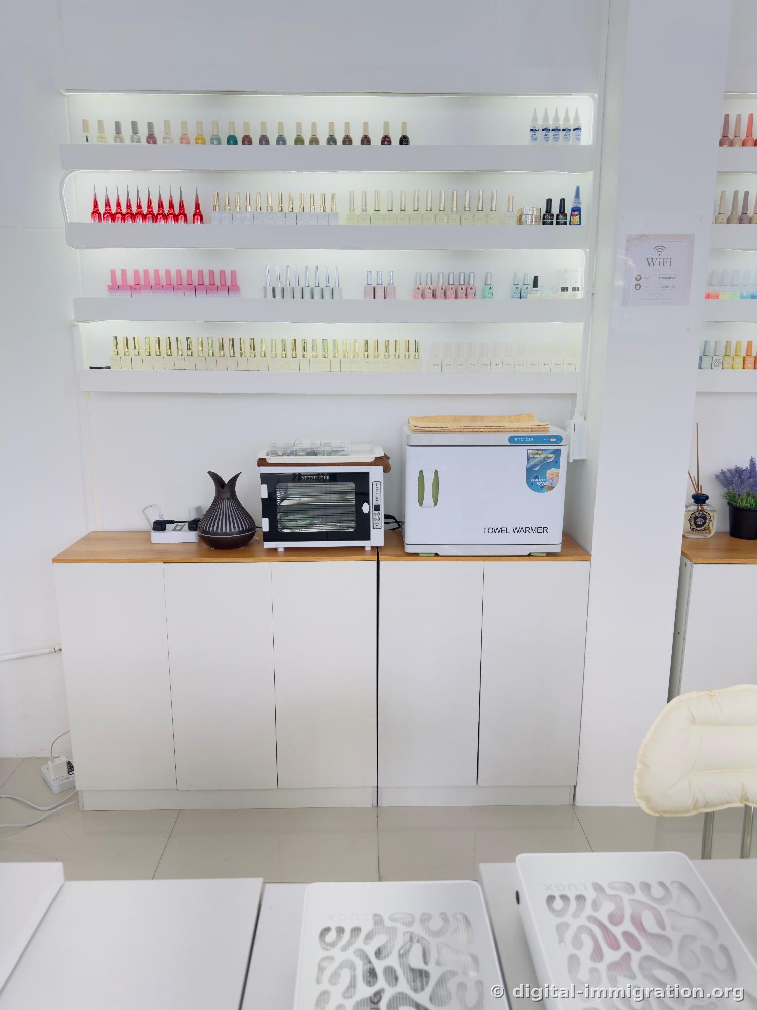 Below the illuminated shelves with nail polish lined up, a sterilizer and towel warmer on the storage cabinet with a diffuser