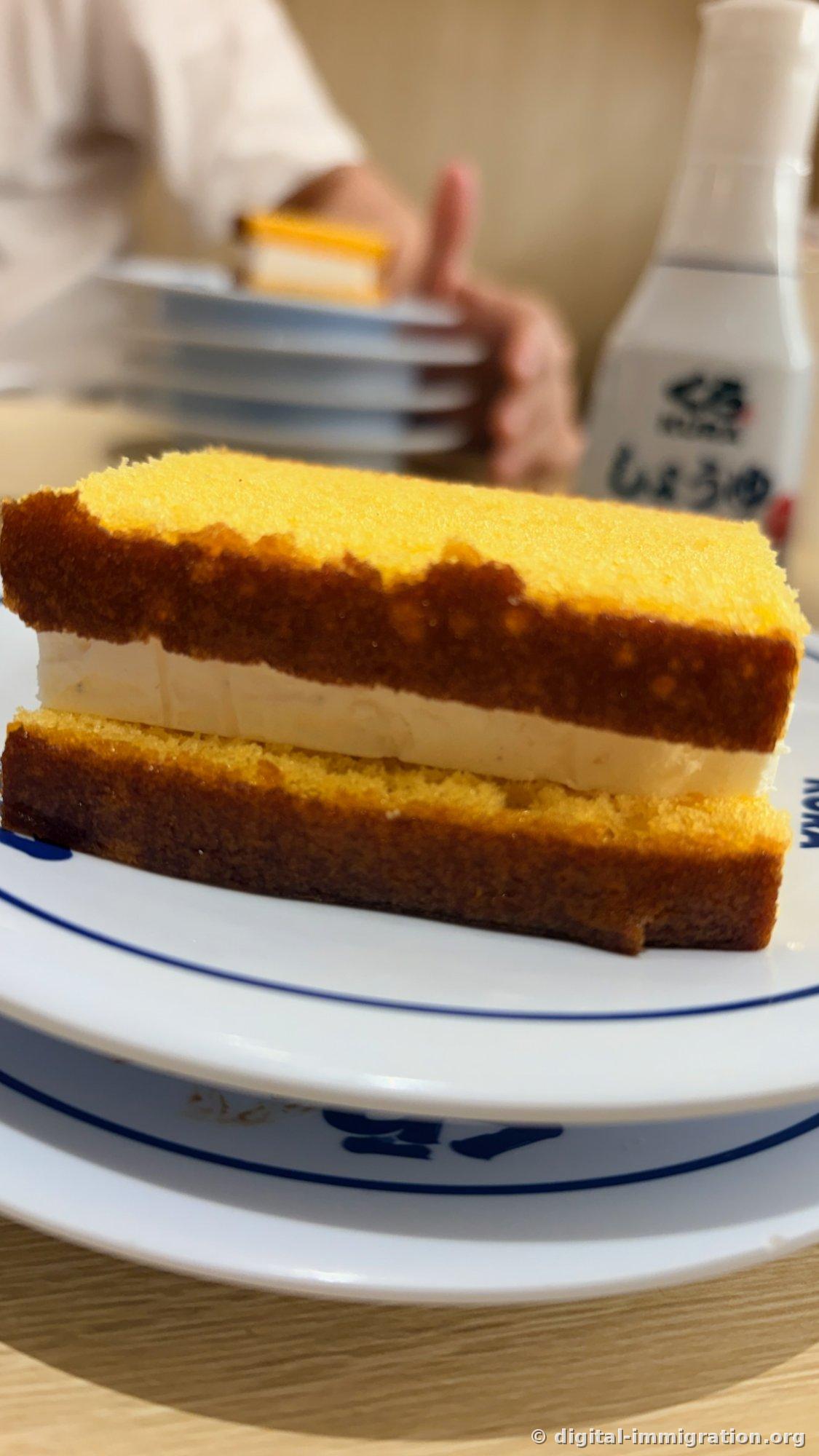 Gâteau Castella vu de côté sur une assiette. Deux tranches de castella jaune avec une épaisse couche de crème entre elles et la tour d'assiettes et la bouteille de sauce soja à l'arrière