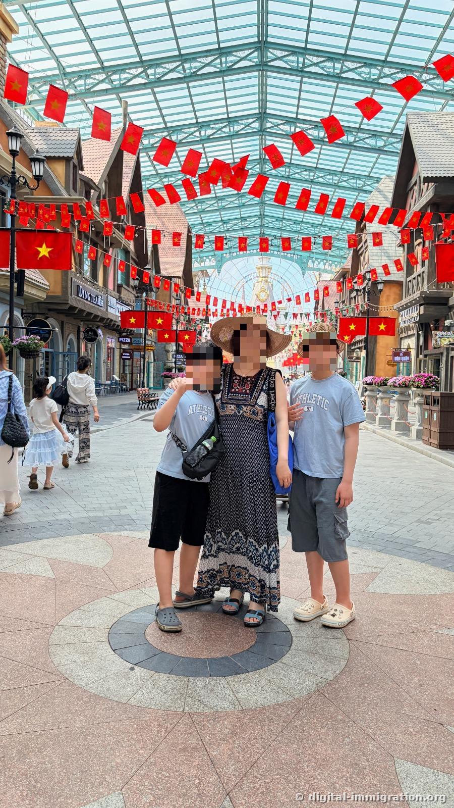 A family standing in an indoor European-style shopping street under a glass ceiling, with Vietnamese flag garlands strung in layers