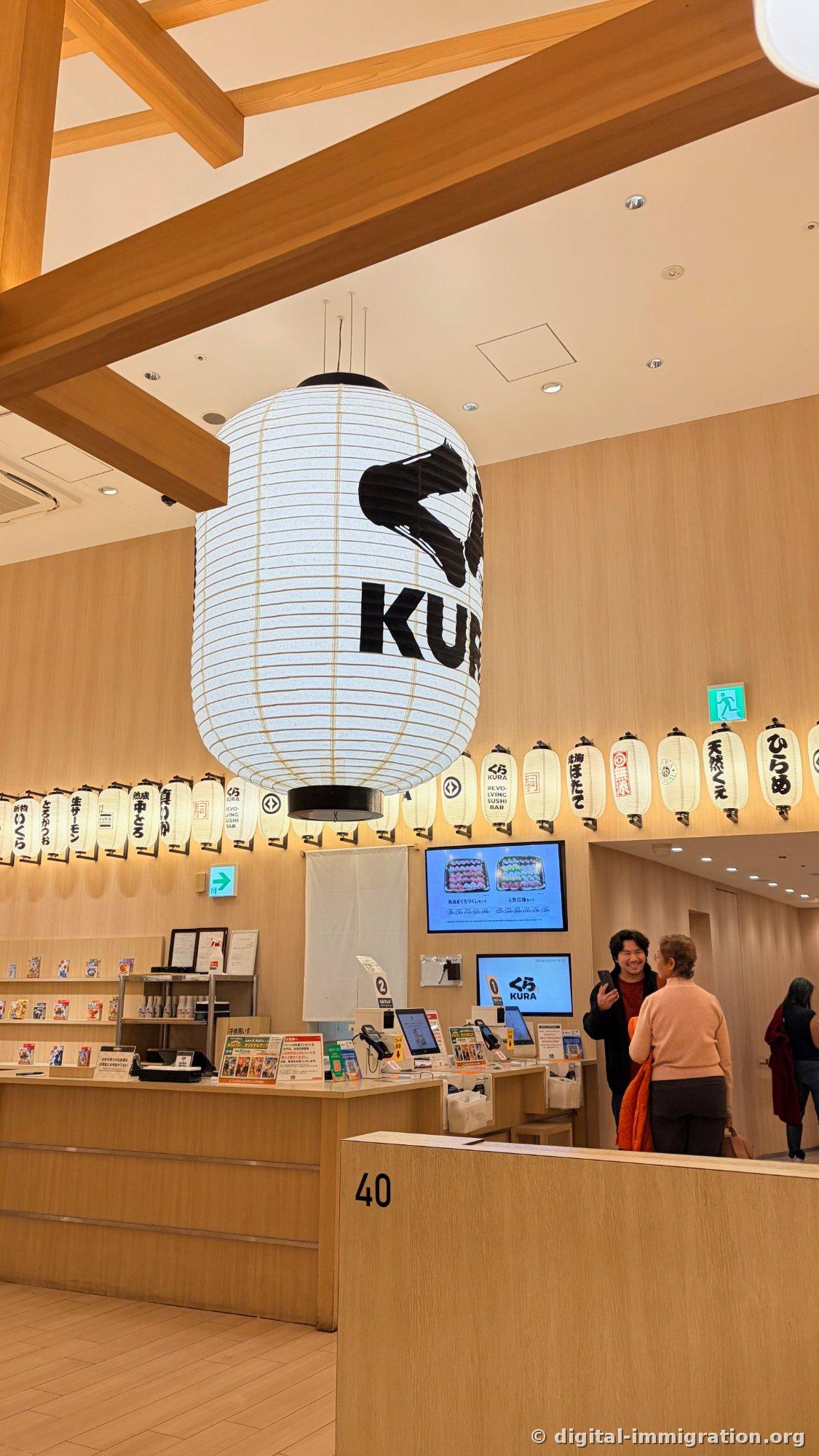 La grande lanterne KURA suspendue au plafond de l'entrée de la succursale Asakusa de Kura Sushi avec les petites lanternes portant les noms de plats, et la caisse de réservation en dessous au comptoir d'accueil